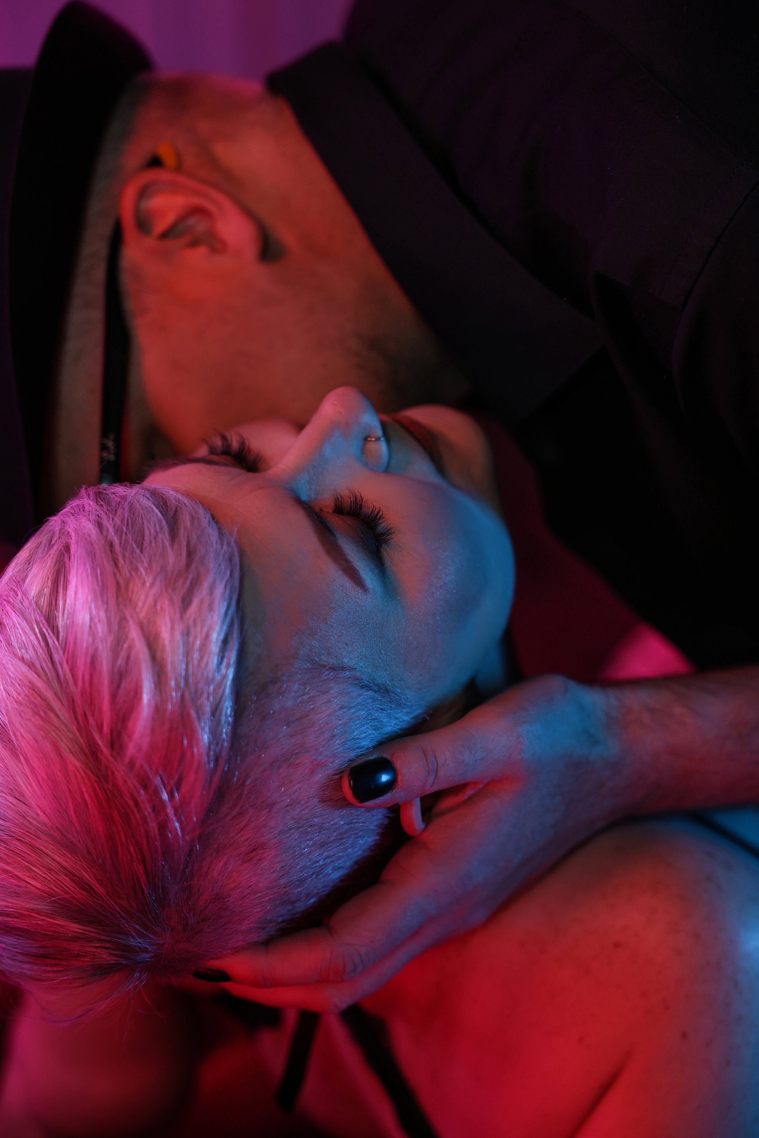 Neon-lit boudoir image of a man in a black hat and glasses embracing a woman with short white hair and kissing her neck