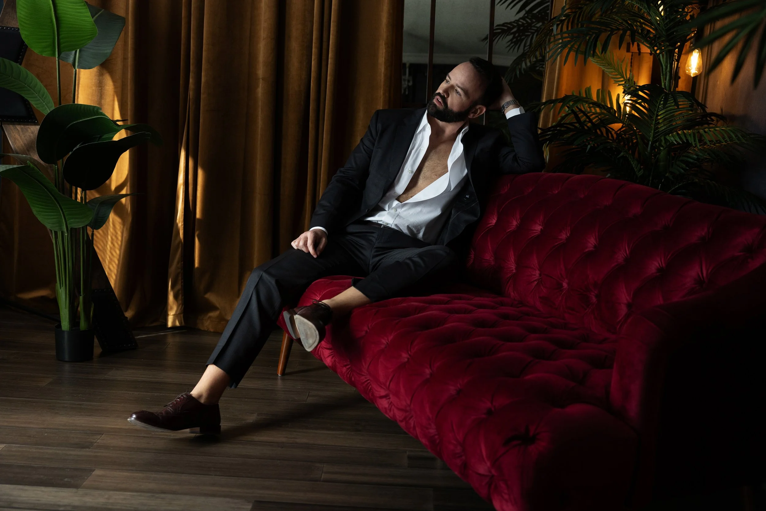Bearded man in dark gray suit lounging on red velvet couch during men’s boudoir session