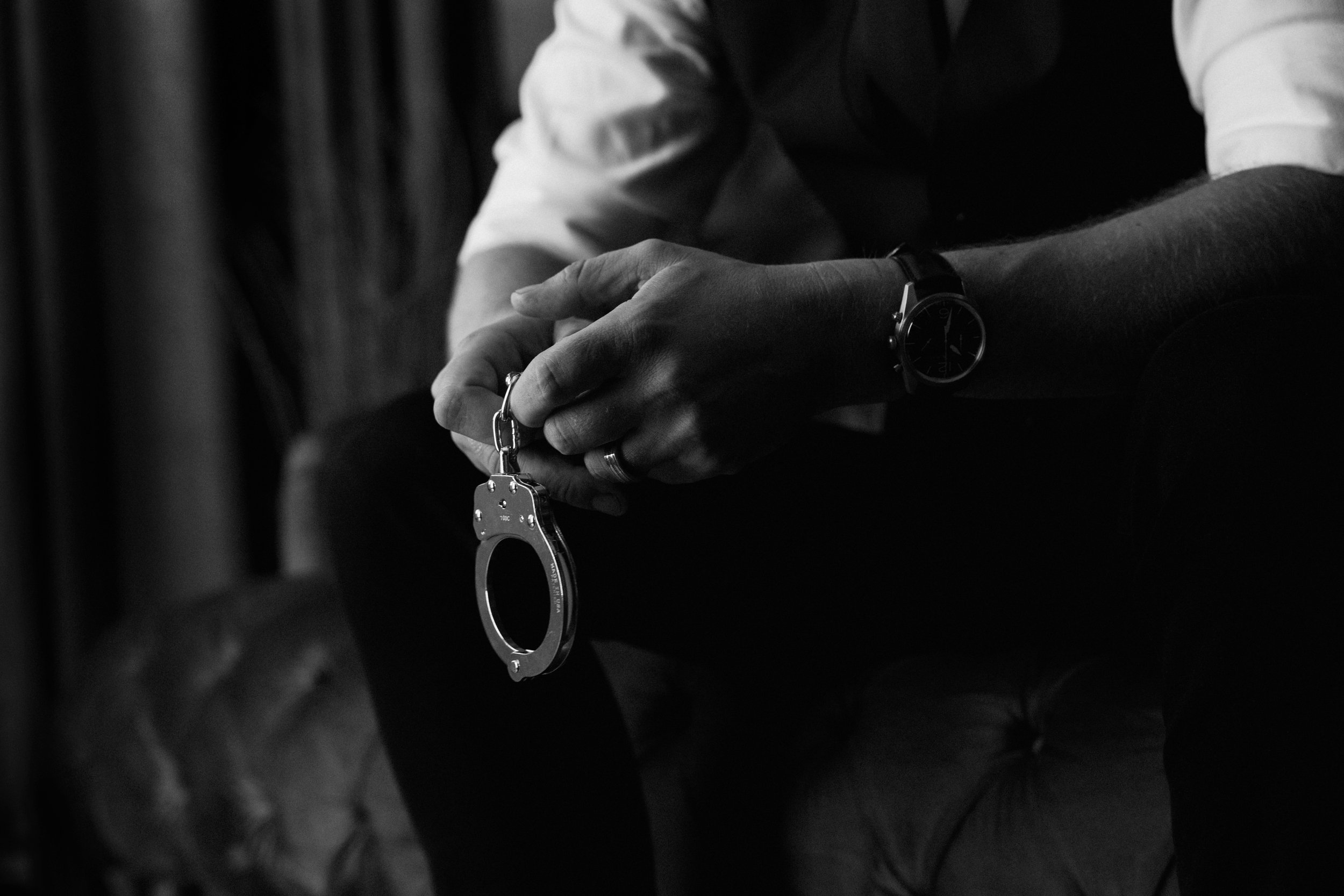 Edgy black and white boudoir portrait of a suited man holding handcuffs while seated on a couch