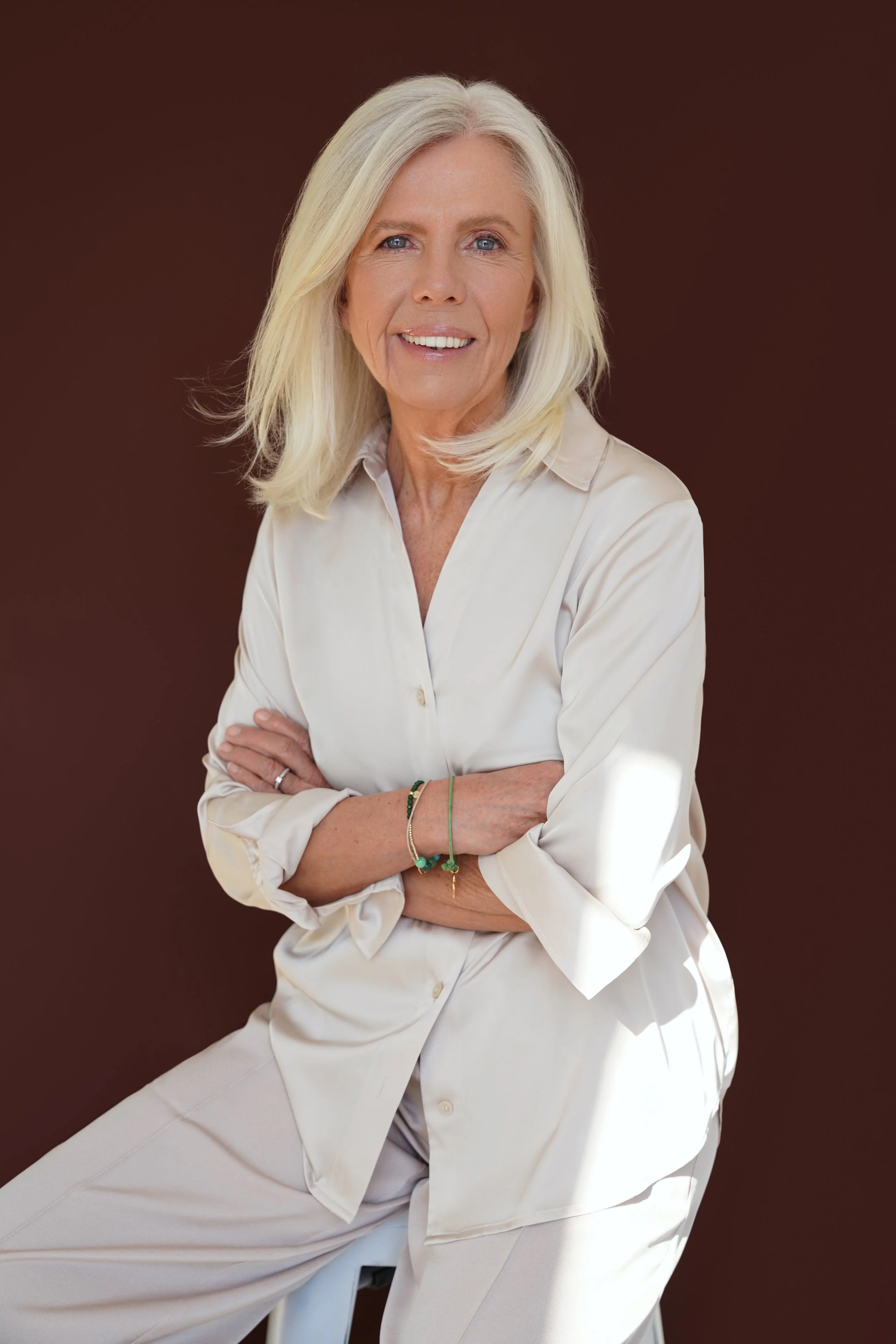 Porträt einer älteren Frau mit langen blonden Haaren, die lächelnd in die Kamera schaut, in einem weißen Outfit, vor einem braunen Hintergrund.