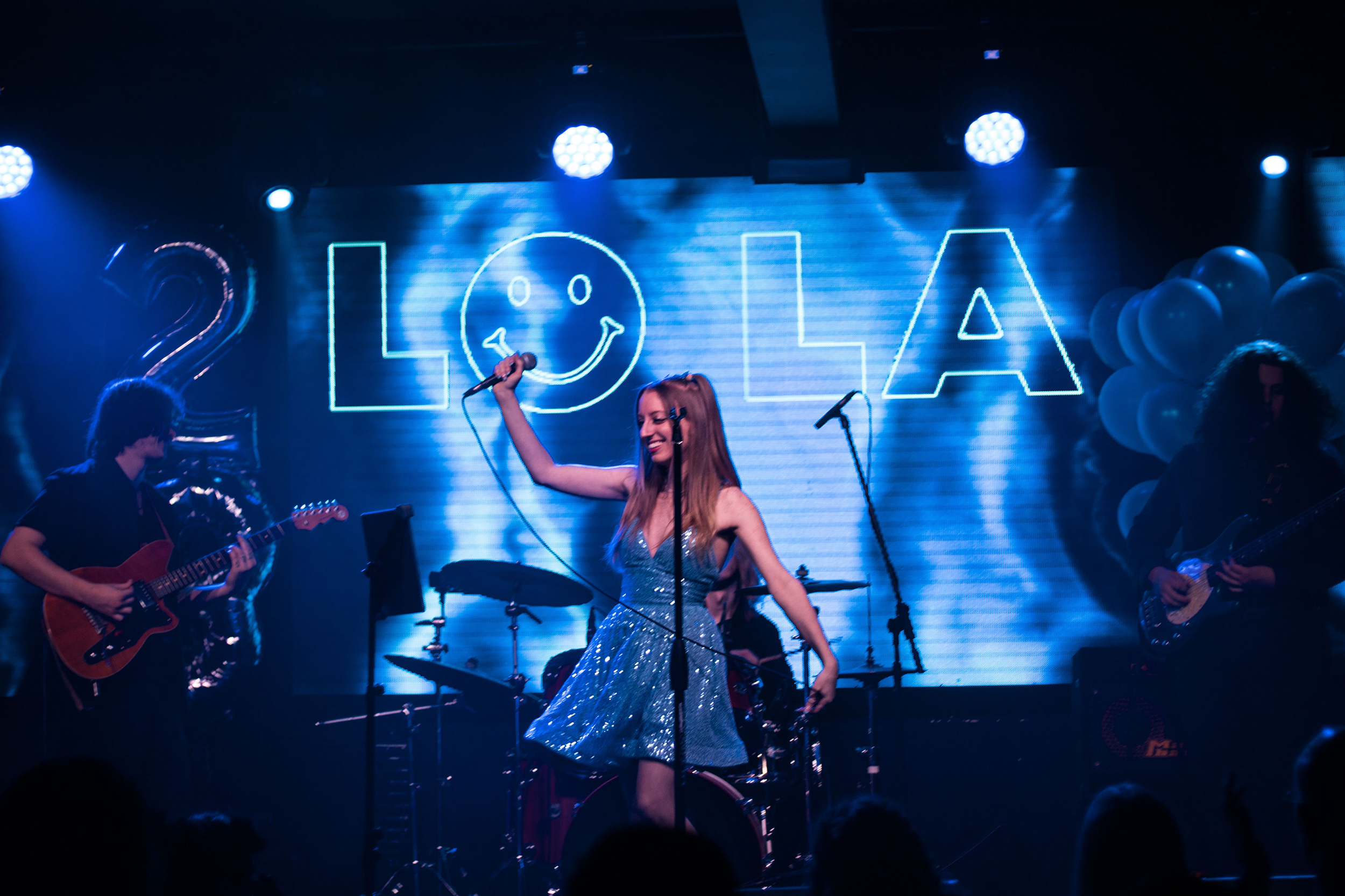 Lola Violet singing and dancing on stage with band members playing guitars in the background, decorated with blue lights and a backdrop that spells out 'LO LA' with a smiley face in the 'O' and balloons on the right.