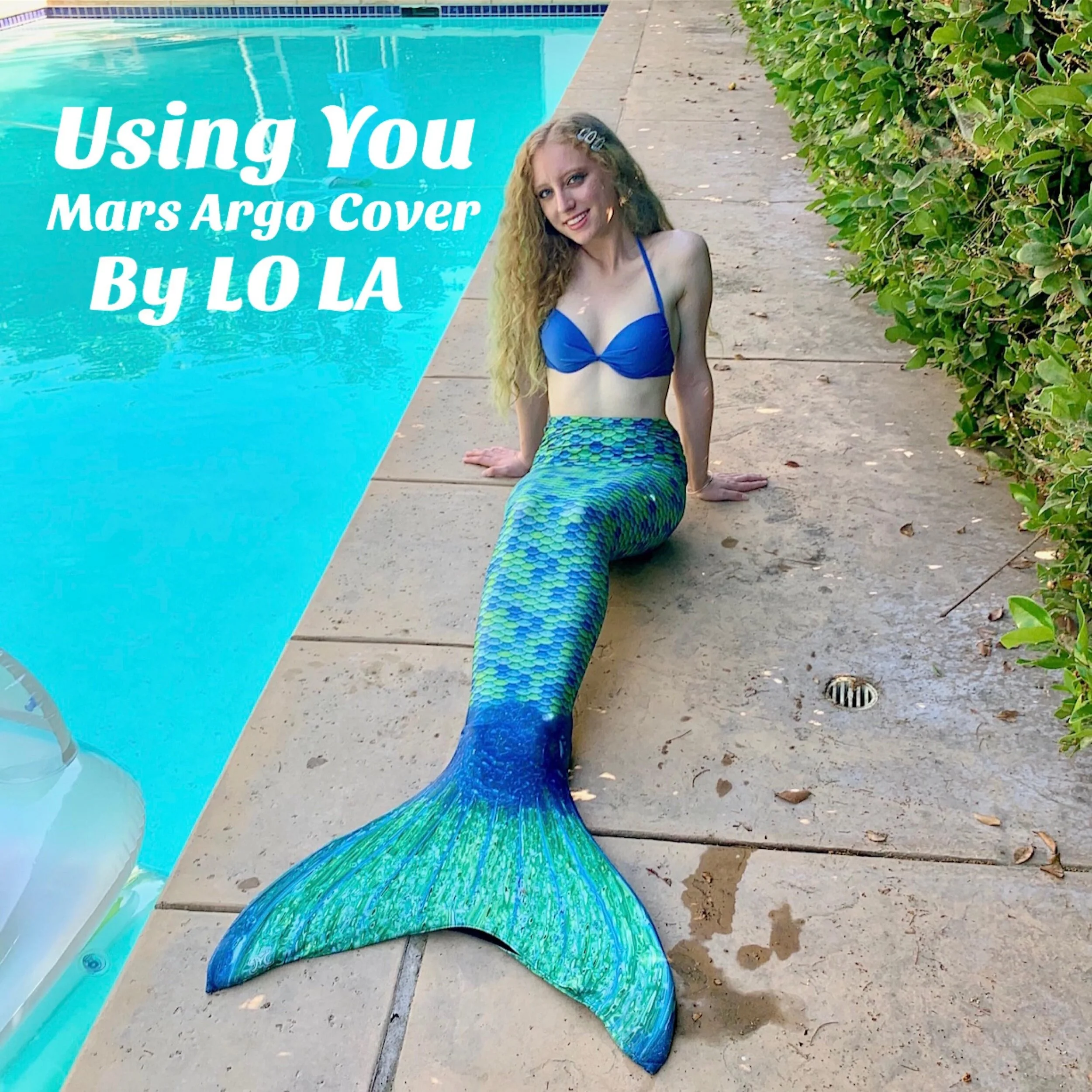Lola Violet dressed as a mermaid with a blue top, colorful tail, and long curly hair, sitting on a poolside concrete surface with a hedge in the background.