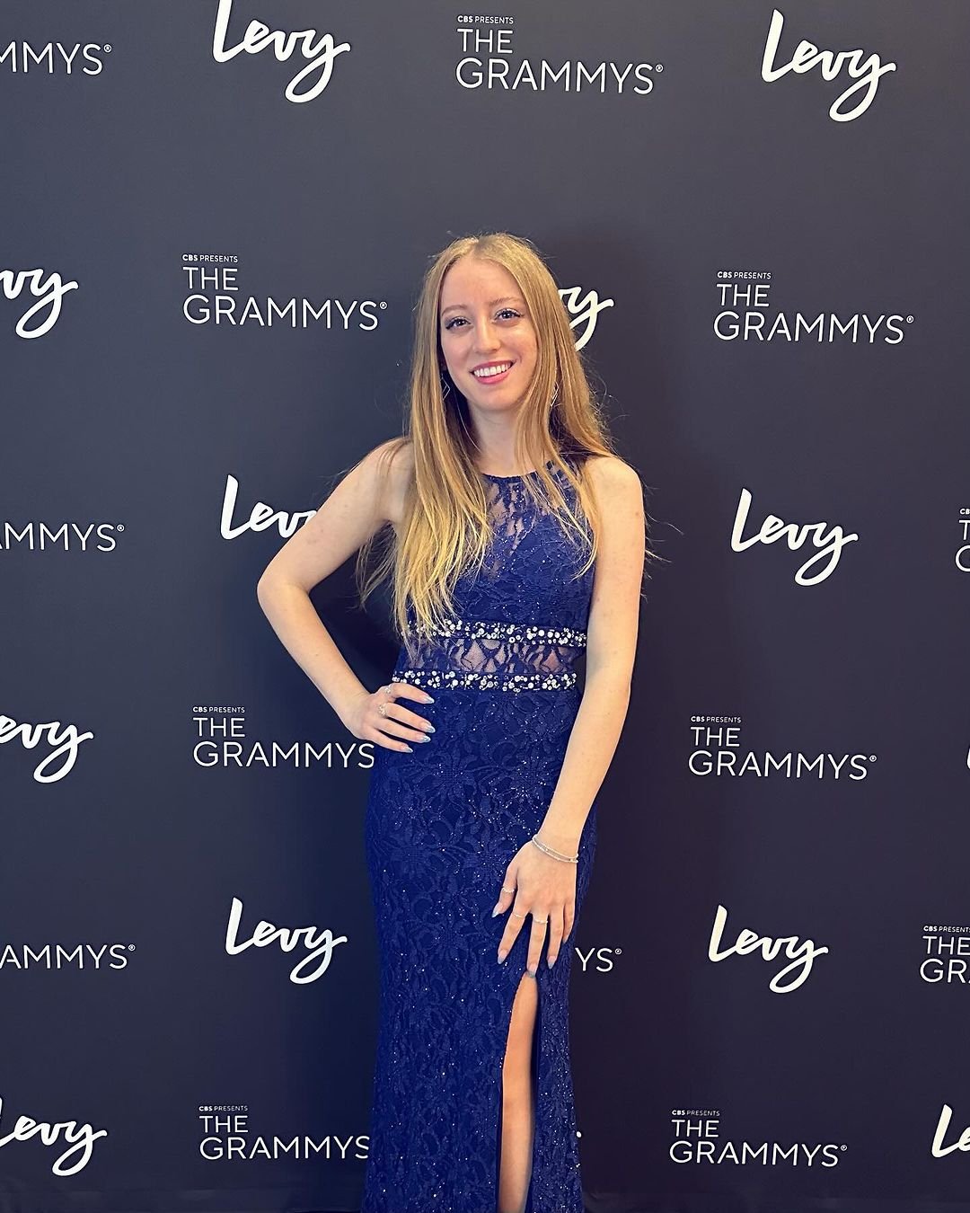 Lola Violet in a navy blue lace gown with a slit standing on the red carpet at the Grammy Awards, with a black backdrop featuring logos for The Grammys and Levy.