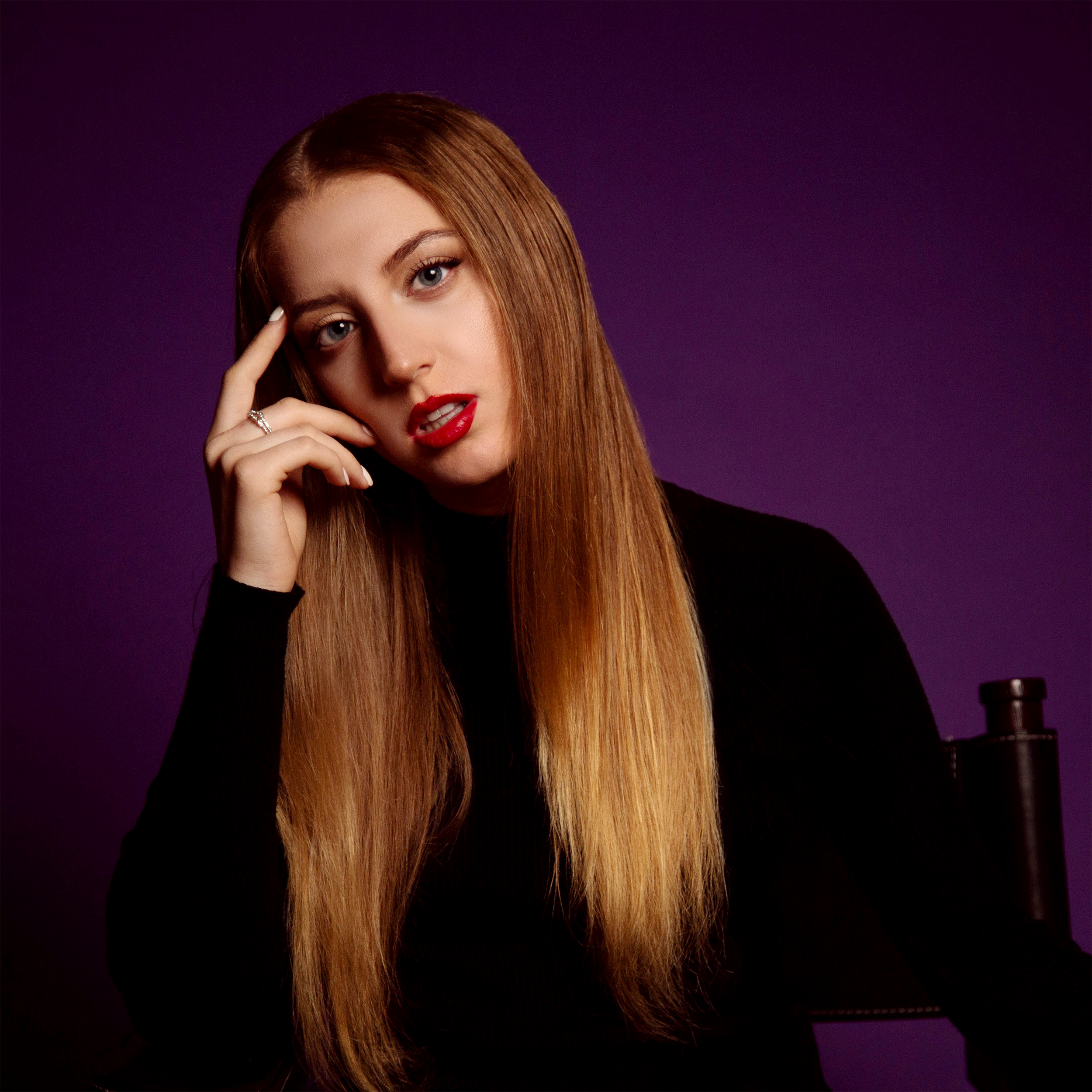 Lola Violet with long red hair, wearing a black top, posing against a purple background. She has makeup and is touching her face with her right hand.