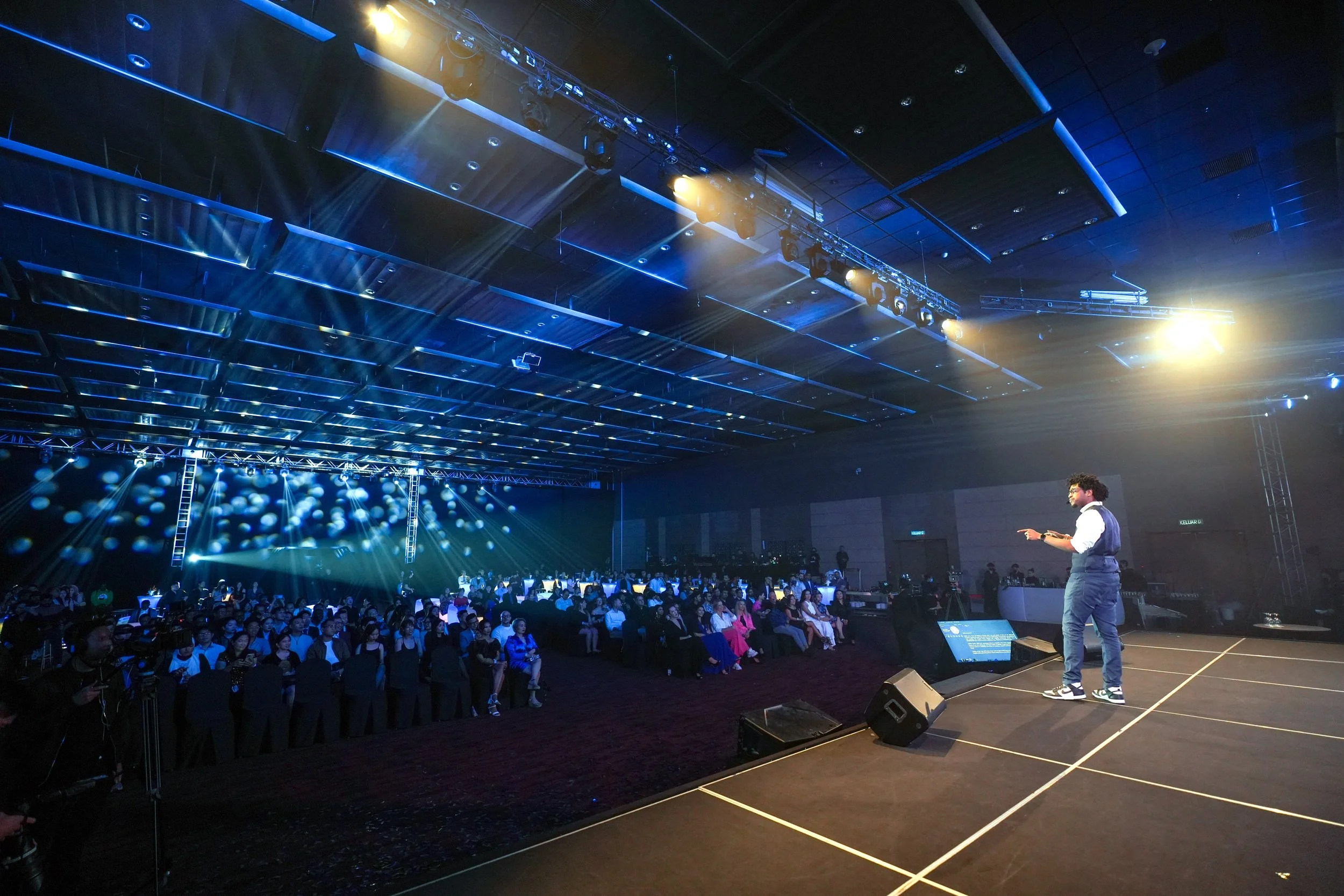 A man with curly hair, glasses, and a goatee, wearing a white shirt with rolled-up sleeves, a dark vest, and dark pants, is standing on stage, smiling and pointing upwards with his right hand. Behind him is a large screen with partial text and images of people, indicating he might be speaking at a conference or seminar.