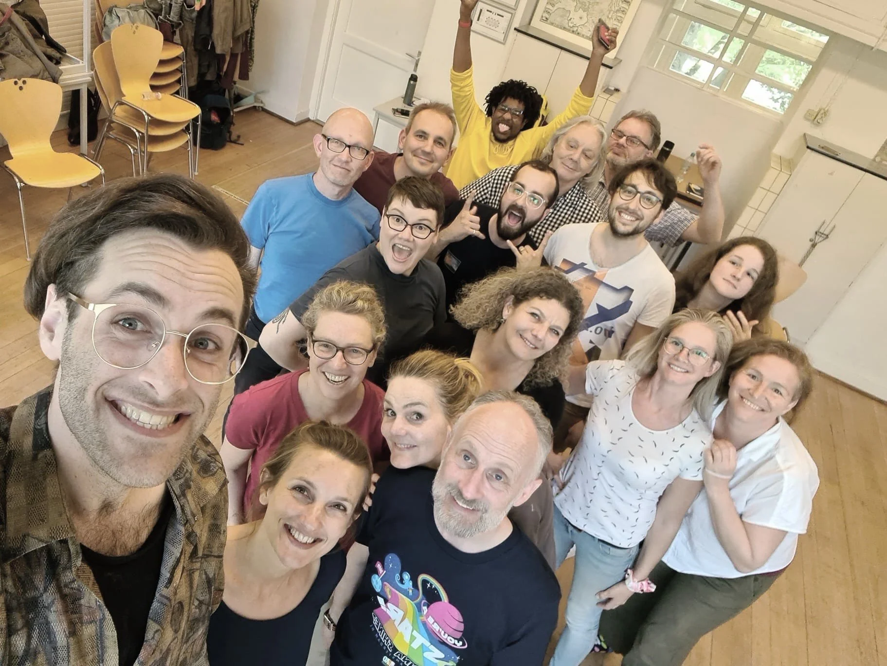 Group of fifteen smiling and cheering people gathered in a room, taking a selfie. The room has wooden floors, white walls, and several chairs stacked in the corner.