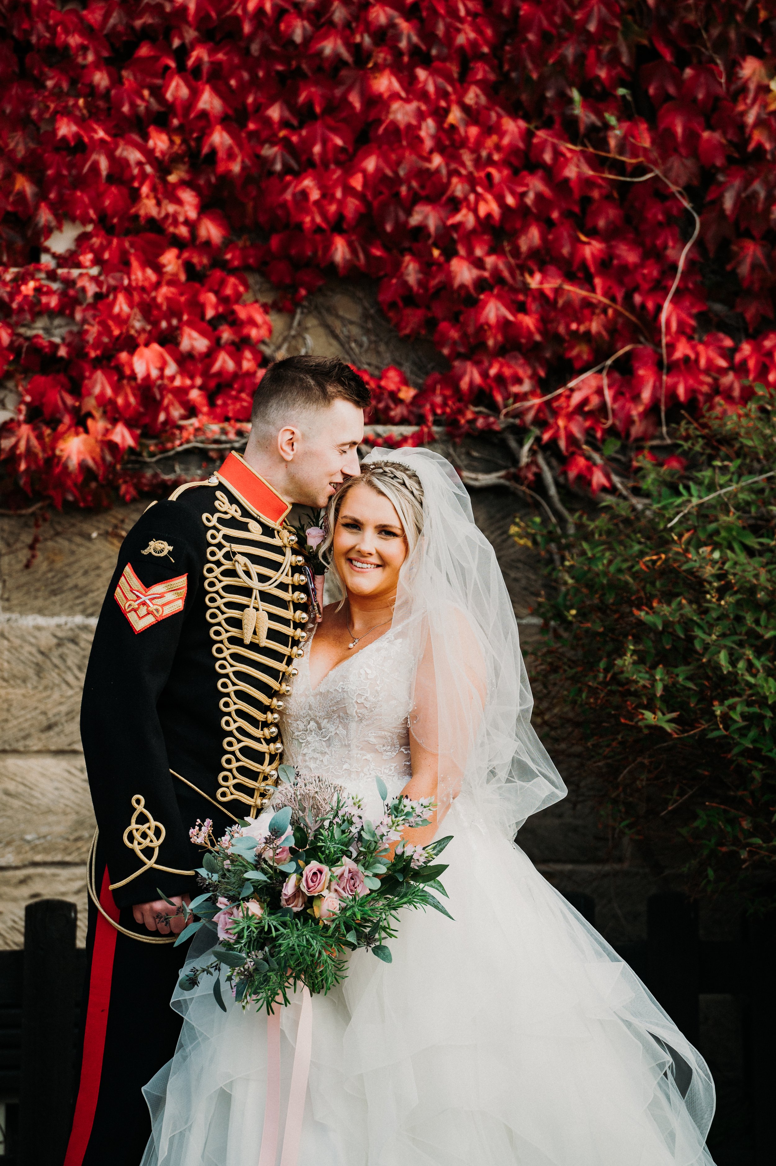 The groom kisses the bride's head in front of some bright red ivy leaves that cling to the walls at Sneaton Castle, Whitby.