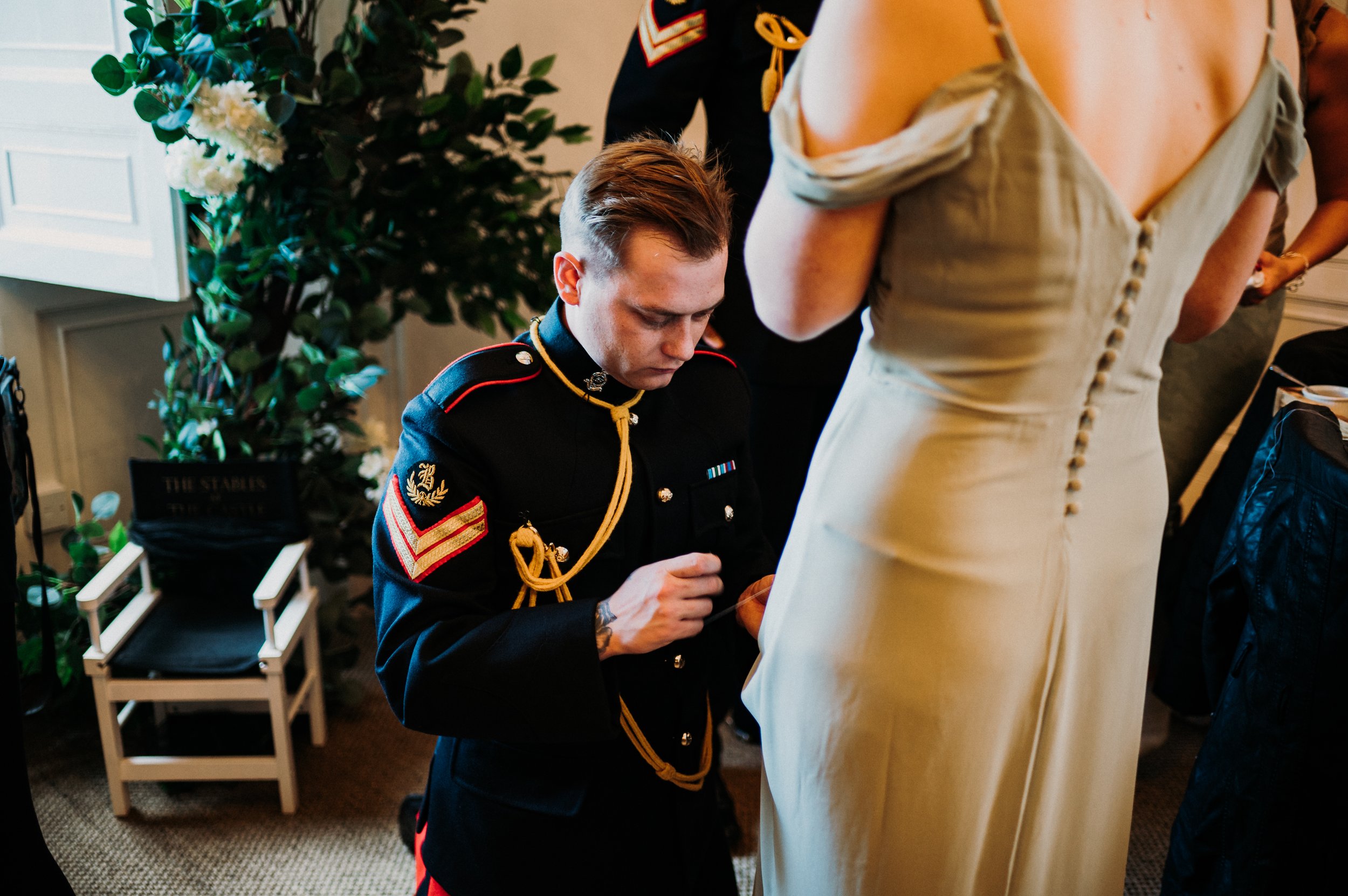 A soldier and master tailor in full uniform helps a bridesmaid with her dress by putting a few stitches in.