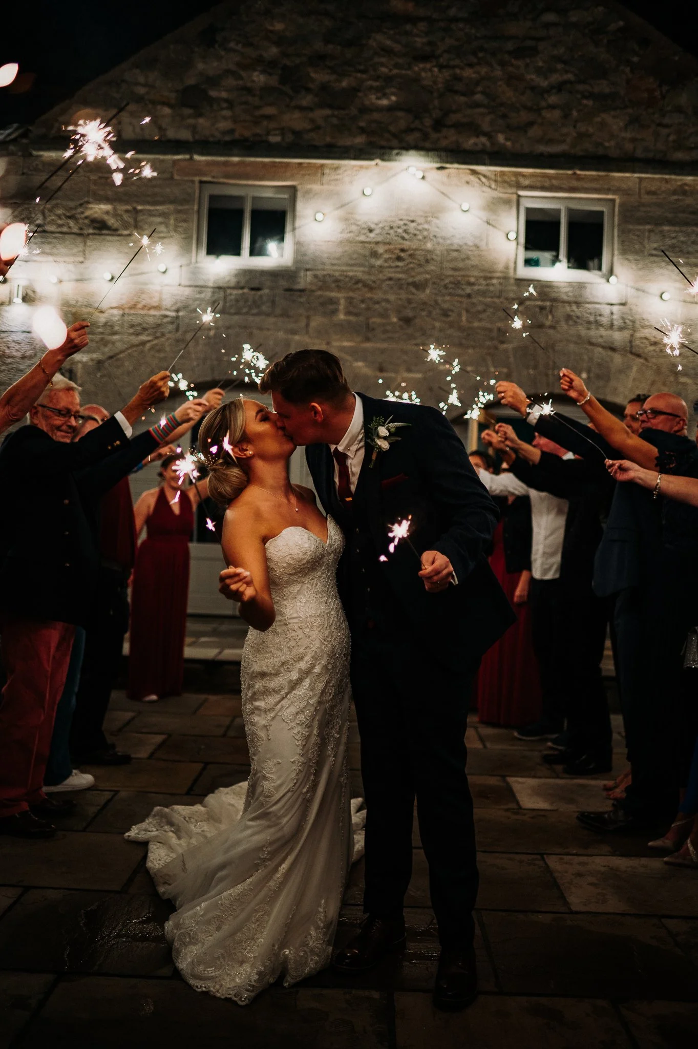 The bride and groom kiss while their guests stand around them with sparklers in the outdoor patio area at Doxford Barns, Northumberland.