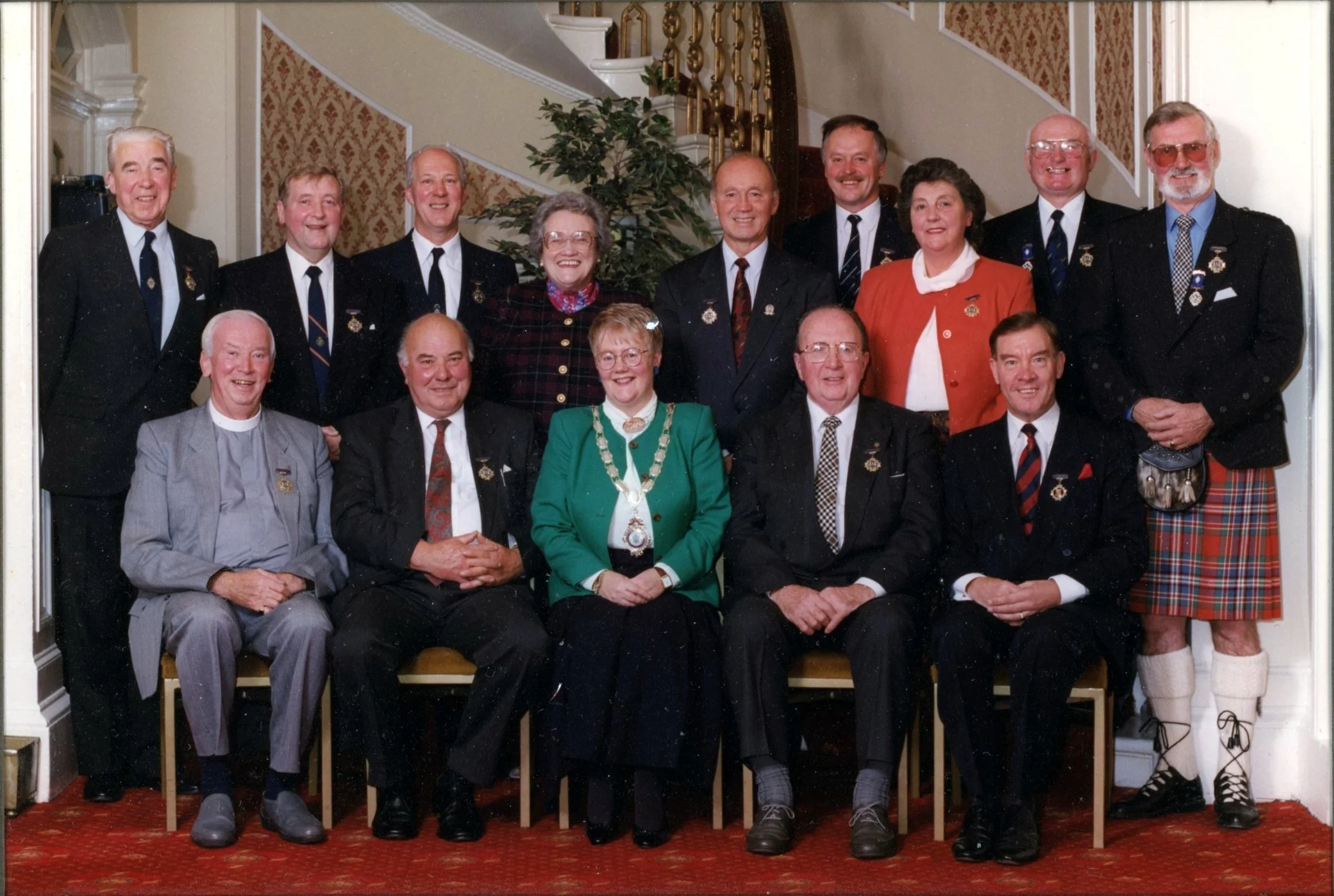 Group of 15 men and women posing for a photo indoors, dressed in business or formal attire, some wearing badges or pins, with many wearing glasses. The background shows a staircase and an ornate interior wall, and the group is arranged in two rows with some seated and others standing.