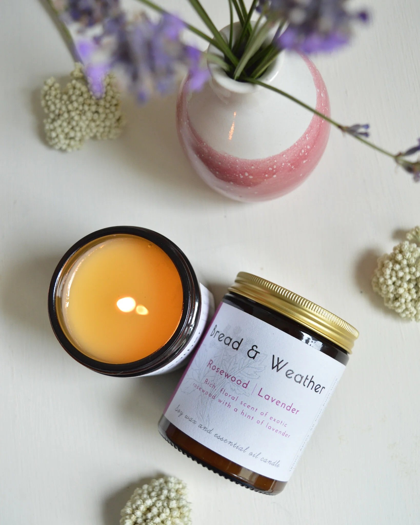 A lit candle, a jar of lavender and rosewood scented oil, a pink and white vase with purple flowers, and small white flowers on a white surface.
