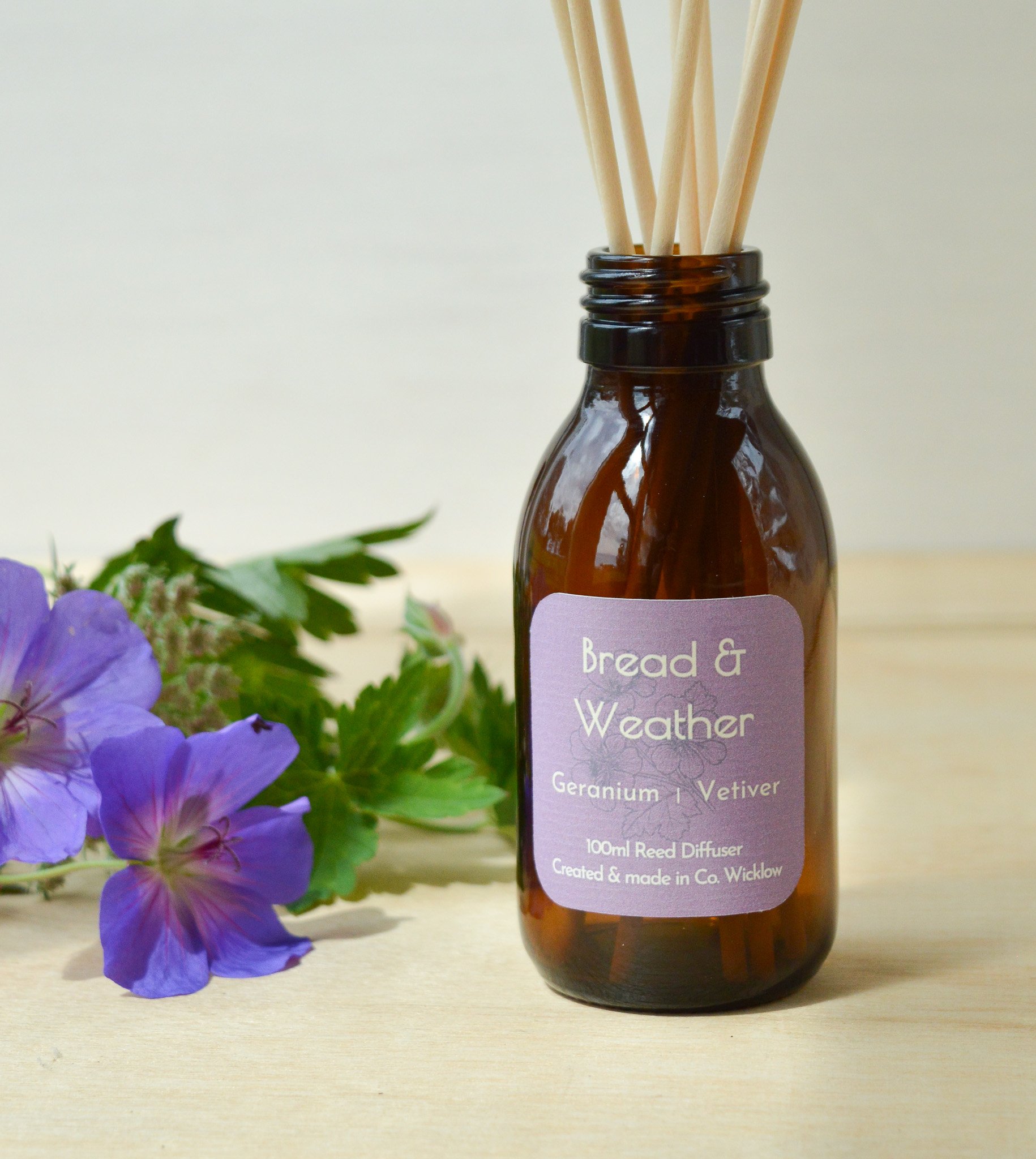 Close-up of Geranium Vetiver diffuser bottle with wild geranium flowers beside it on wooden surface