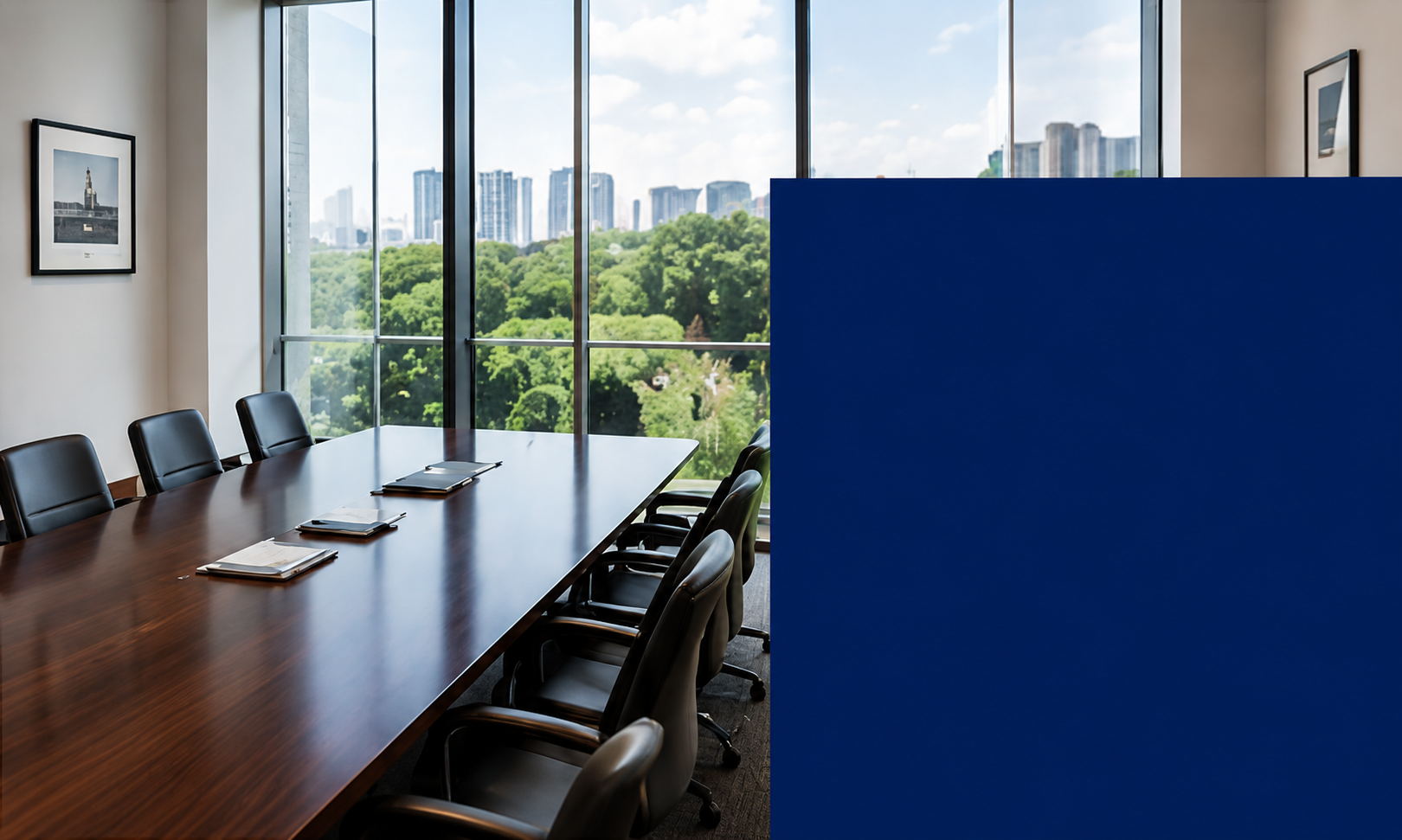 Empty conference room with wooden table, black chairs, large windows overlooking green trees and city skyline, framed pictures on white walls, and a blue partition on the right side.