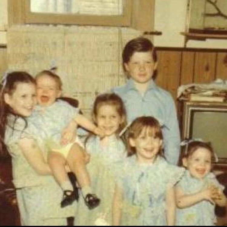 Group of five children and one adult standing in a room with wooden walls and shelves, all smiling.