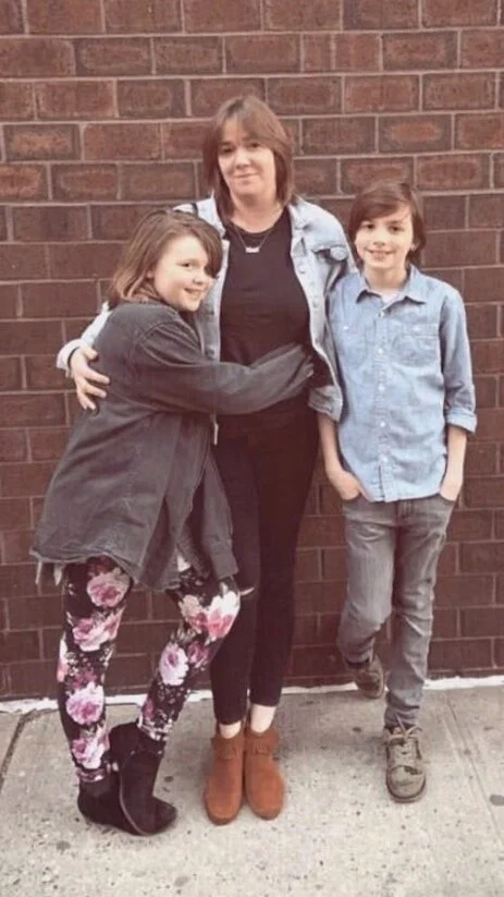 A woman with two children standing in front of a brick wall, with the children hugging her.