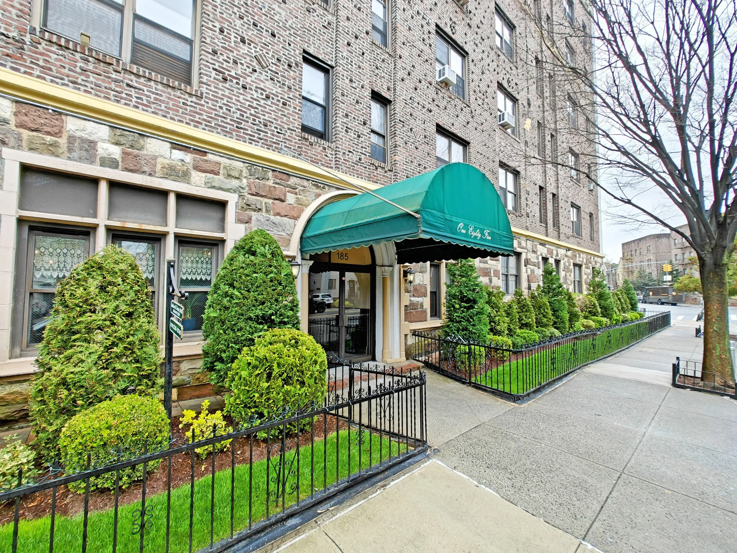 Apartment building with a green awning over the entrance, surrounded by neatly trimmed bushes and a small garden, situated along a city sidewalk on a cloudy day.