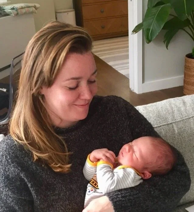 A woman holding a newborn baby in her arms, both smiling at each other in a cozy living room.