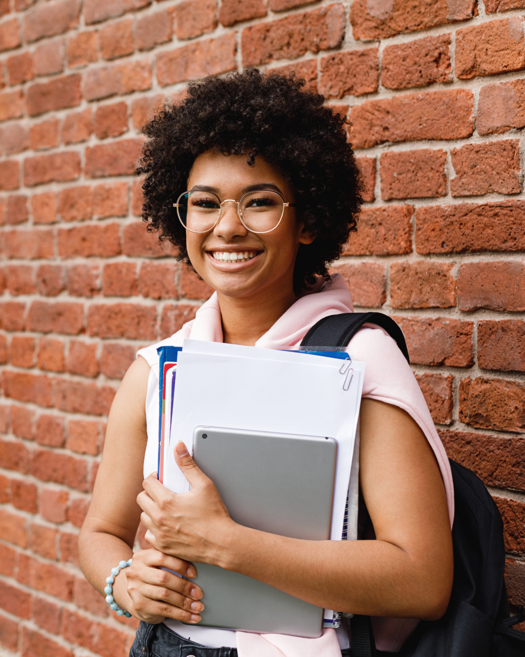 Young woman with glasses, curly hair, smiling, holding a laptop and folders, standing against a brick wall.