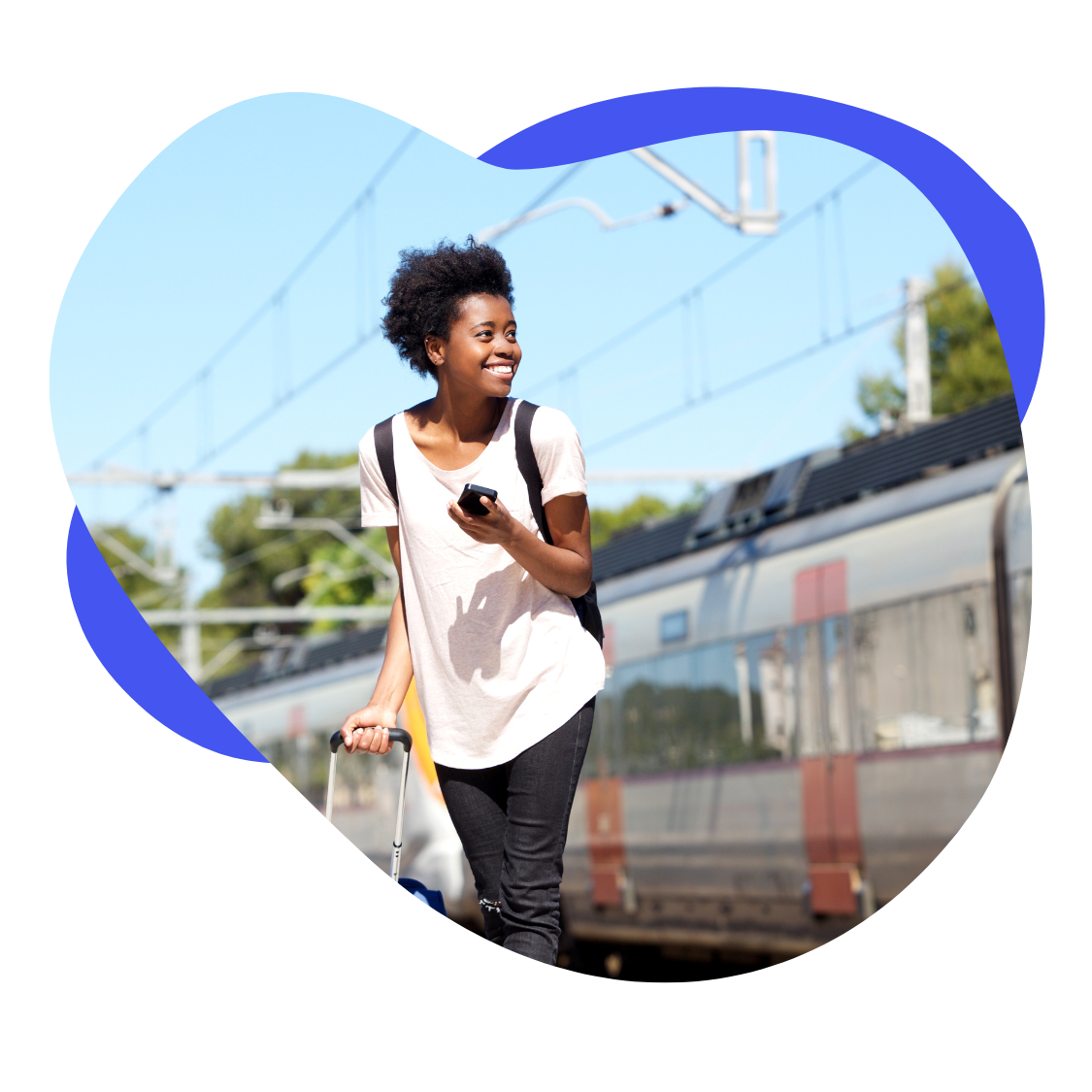 A smiling young woman with a backpack and rolling suitcase walking at a train station, holding a mobile phone.