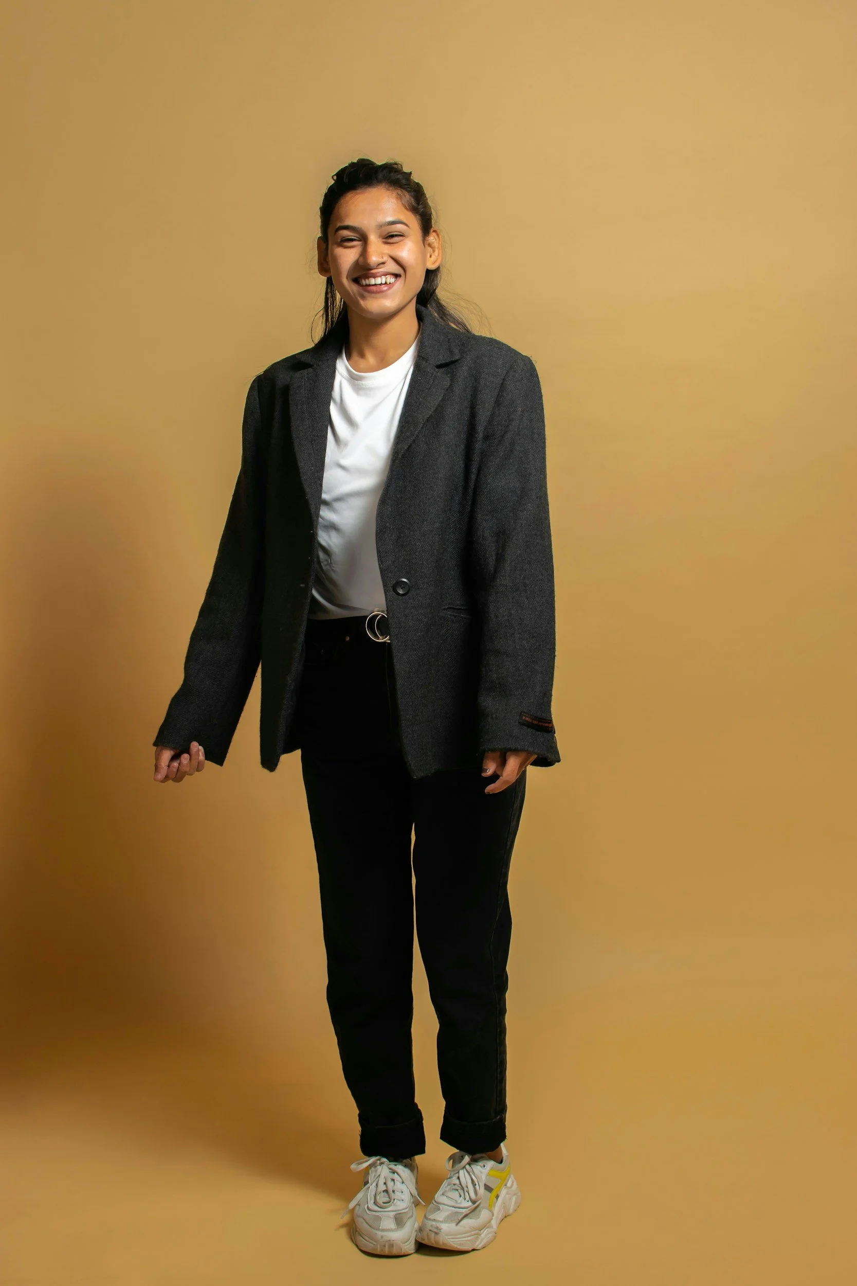 A young woman with dark hair styled in a half-up, half-down look, smiling brightly at the camera, dressed in a dark blazer, white t-shirt, black pants, and white sneakers, standing against a plain yellow background.