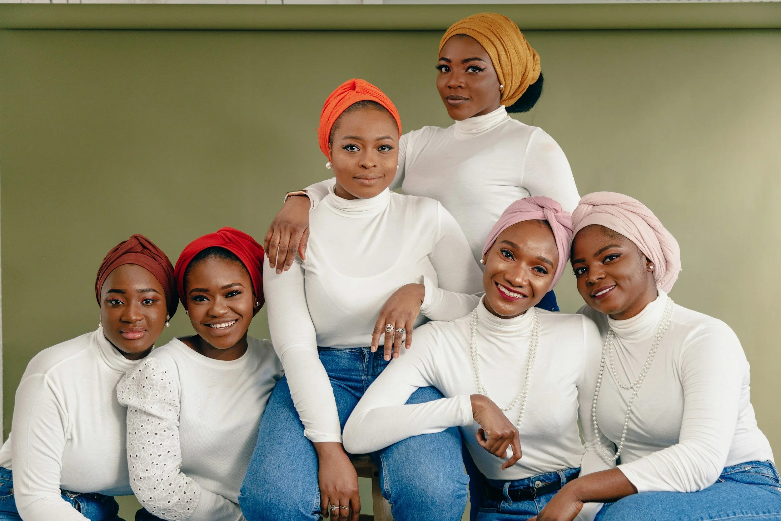 Group of six women wearing white tops, with colorful headscarves, sitting and standing together against a neutral background, smiling and posing for the photo.