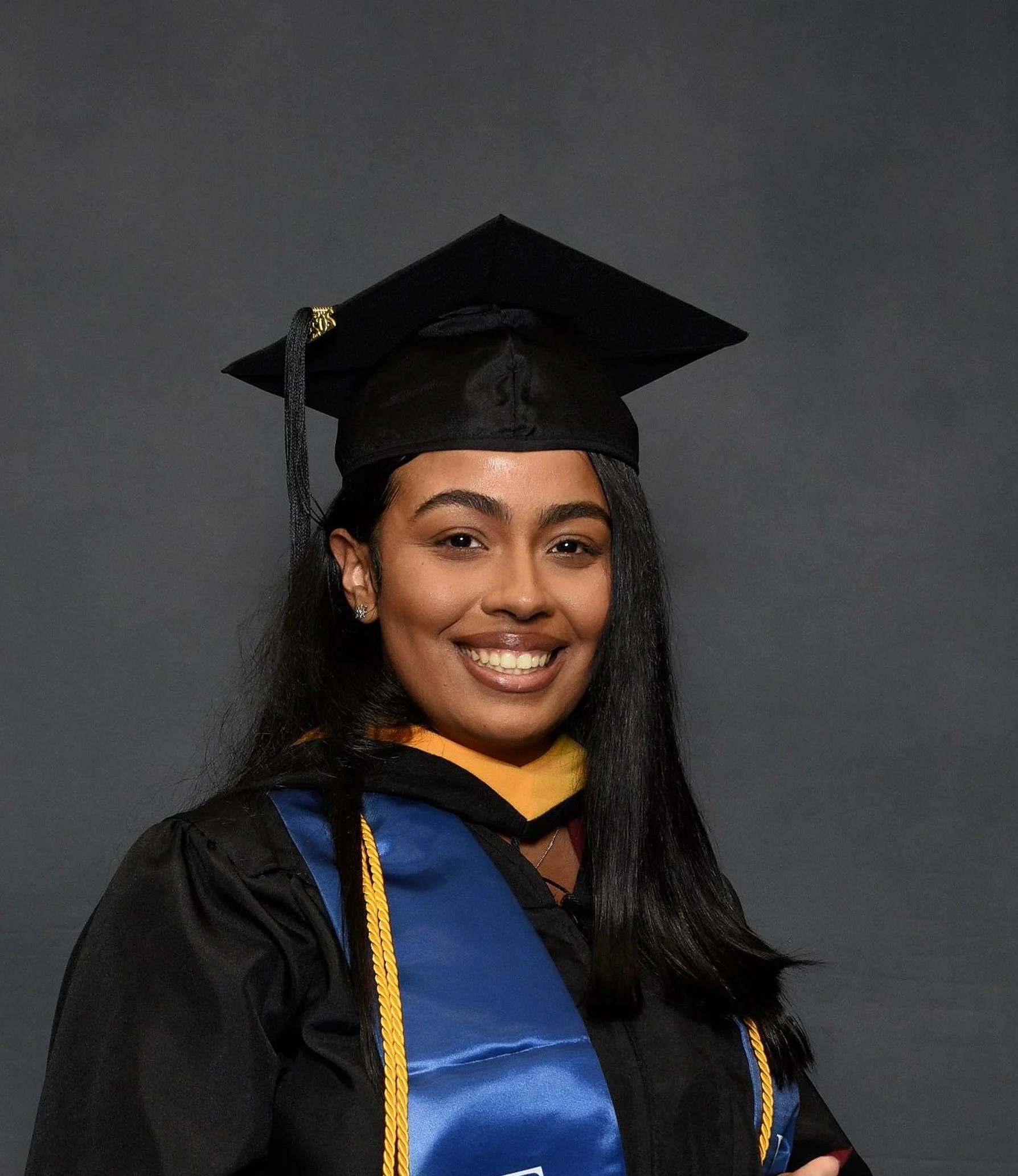 A young woman wearing a graduation cap and gown, smiling, standing against a dark background.