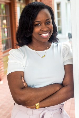 A woman with dark hair in a bob style, smiling, wearing a white top, gold bracelet, and necklace, standing outdoors near a window with a porch in the background.