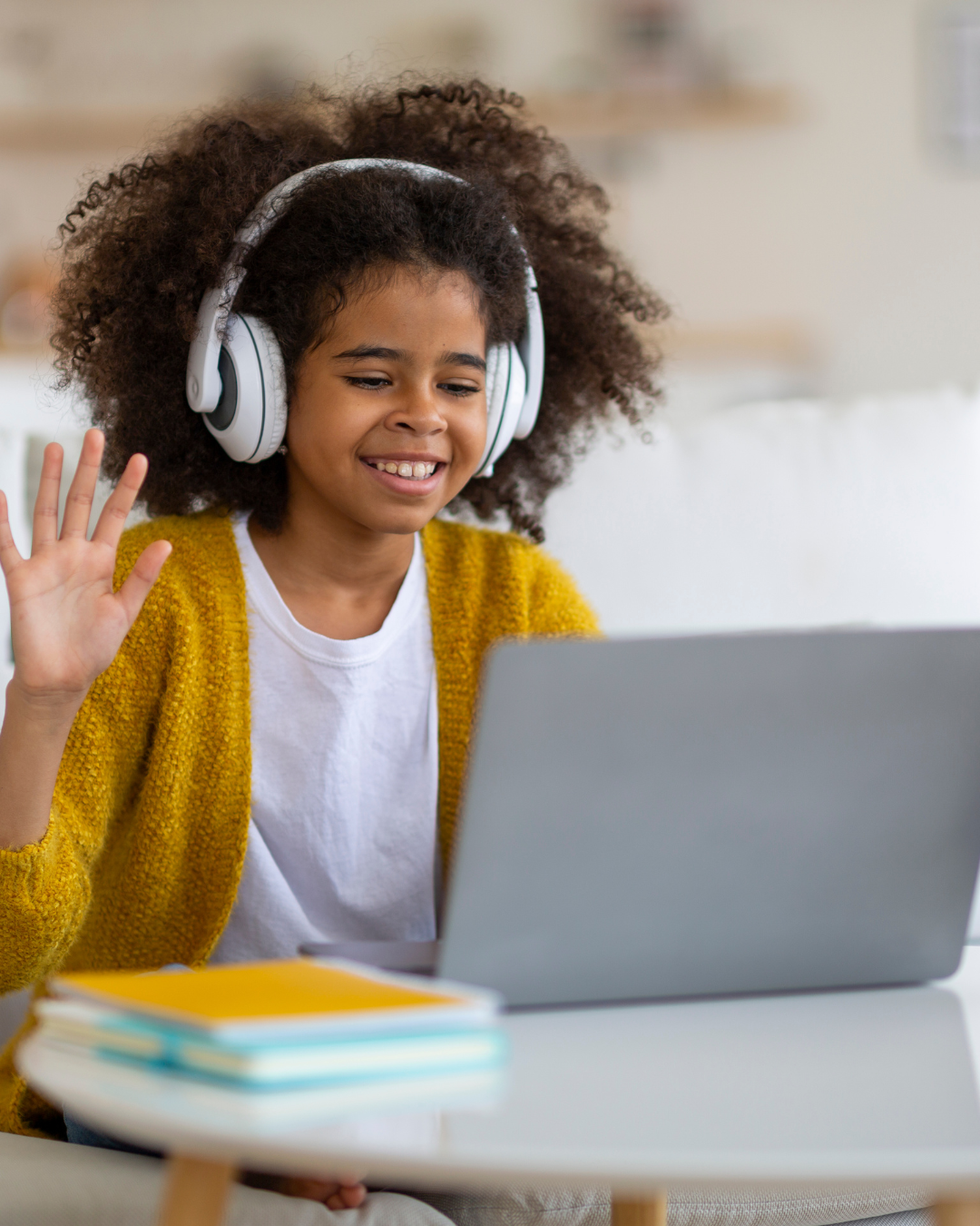 A young girl with curly hair wearing headphones, a white shirt, and a yellow cardigan, smiling and waving at her laptop while sitting at a white table with stacked notebooks.