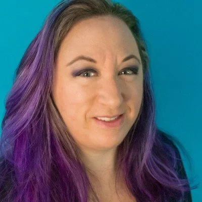 A woman with purple hair smiling at the camera against a blue background.