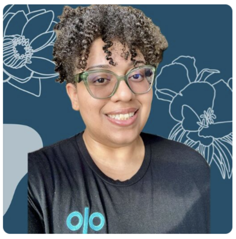 A smiling woman with curly hair and glasses, wearing a black t-shirt with a blue logo, standing in front of a blue background with white floral line art.