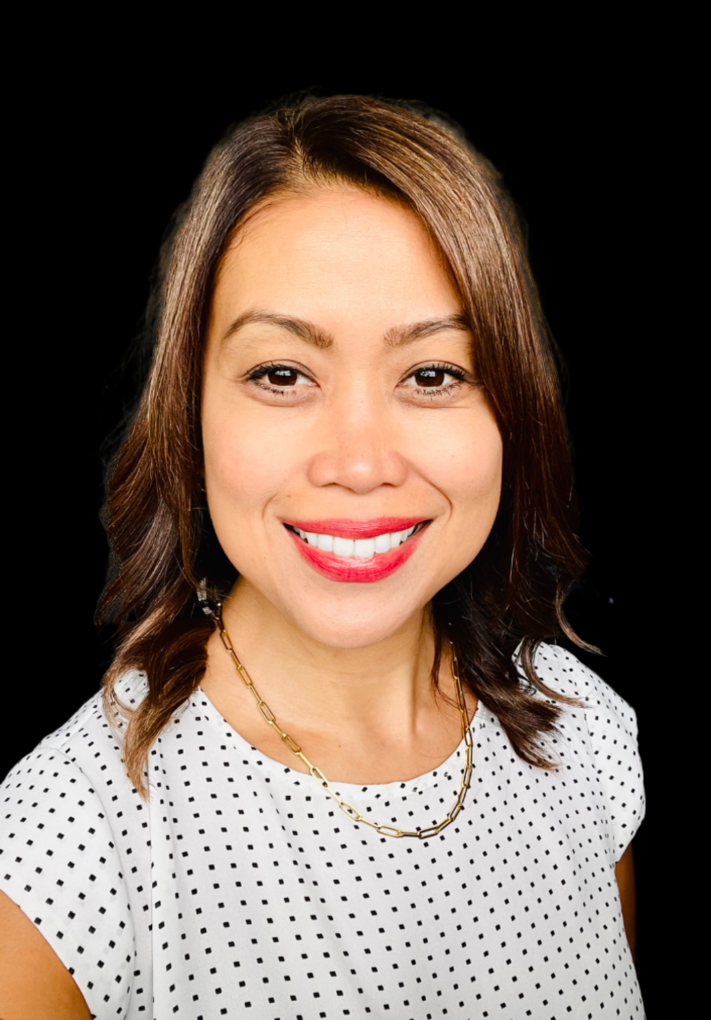 Close-up portrait of a smiling woman with brown wavy hair, wearing a white polka dot blouse and gold chain necklace, against a black background.