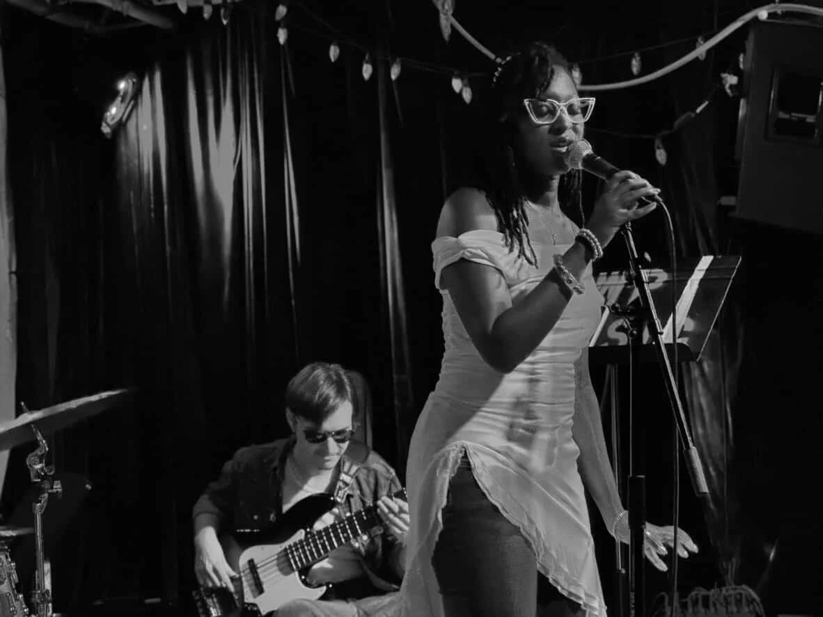 A woman singing into a microphone on stage with a musician playing guitar behind her, set against a dark curtain backdrop.
