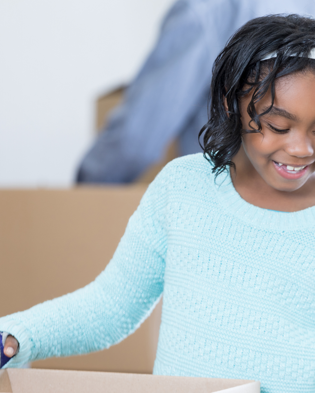 A young girl with dark, curly hair smiling while packing or unpacking a cardboard box.