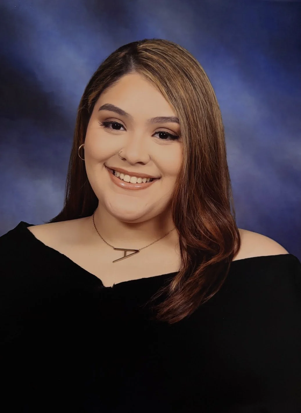 A young woman with long, reddish-brown hair smiling, wearing a black off-shoulder top, a necklace with a letter pendant, hoop earrings, and a nose ring, against a dark blue background.