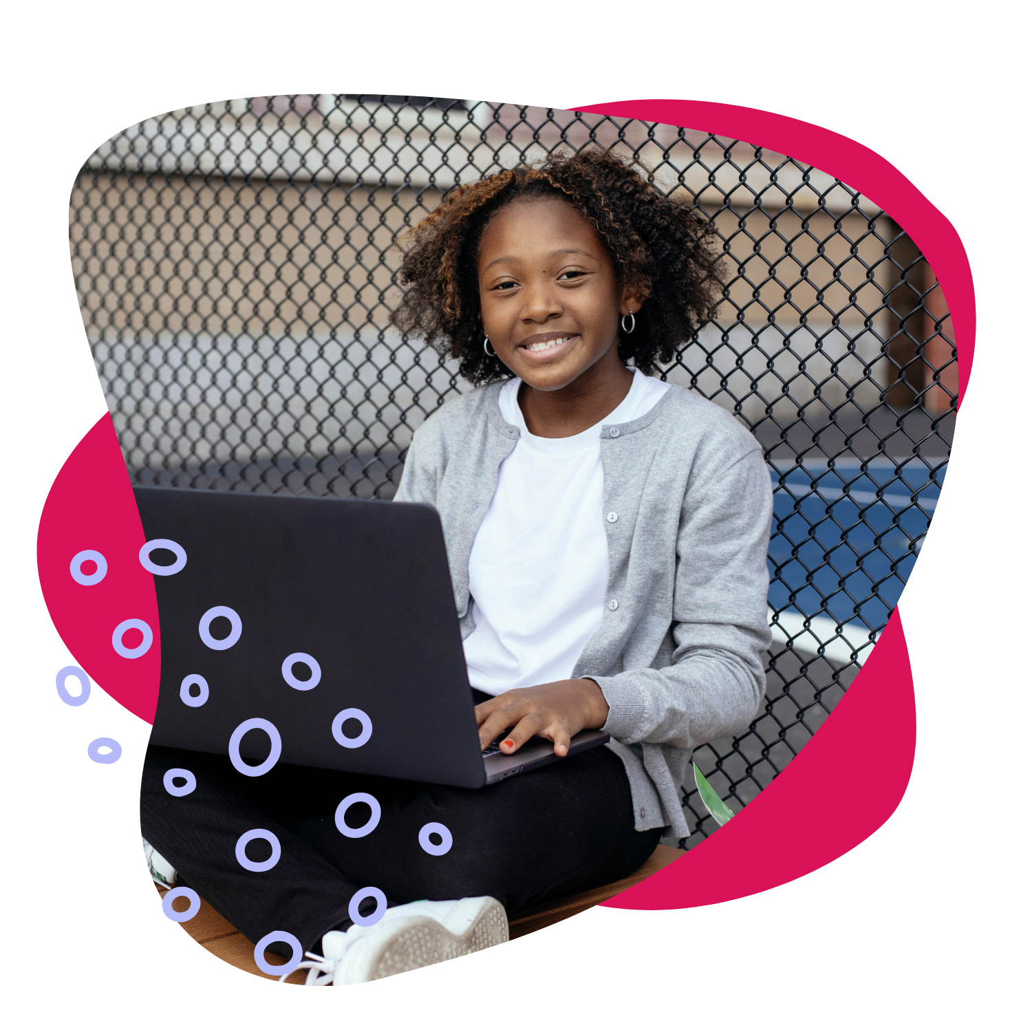 A young girl sitting outdoors in front of a chain-link fence, smiling while using a laptop.
