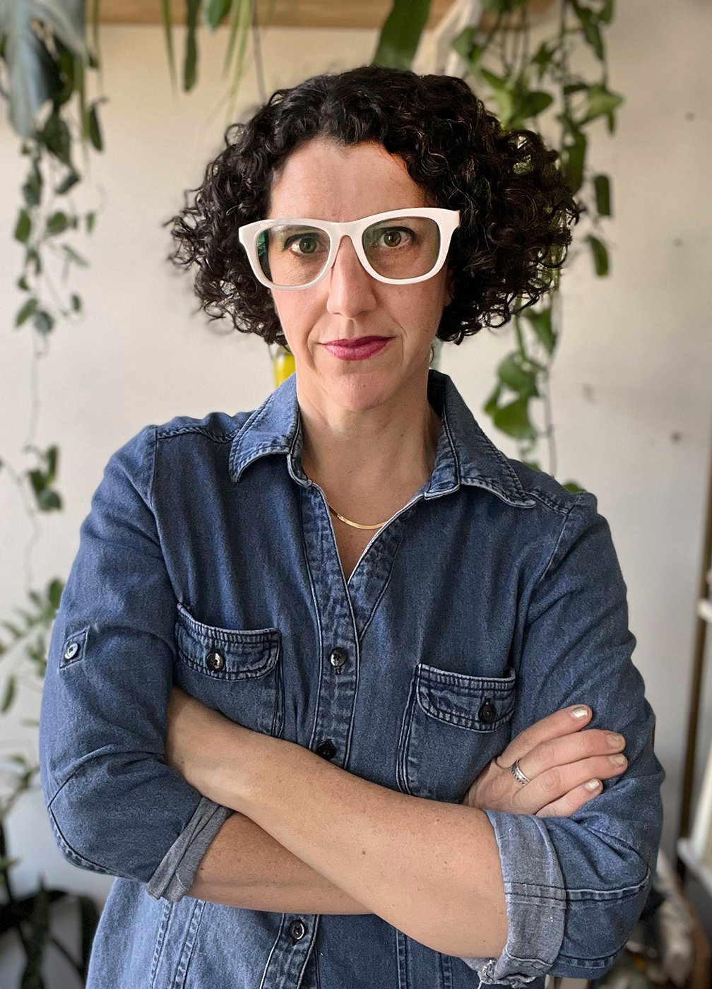 Photograph of Sonya, an artist with brunette curly hair styled in a bob. She is wearing white glasses and a denim work shirt and her arms are crossed.