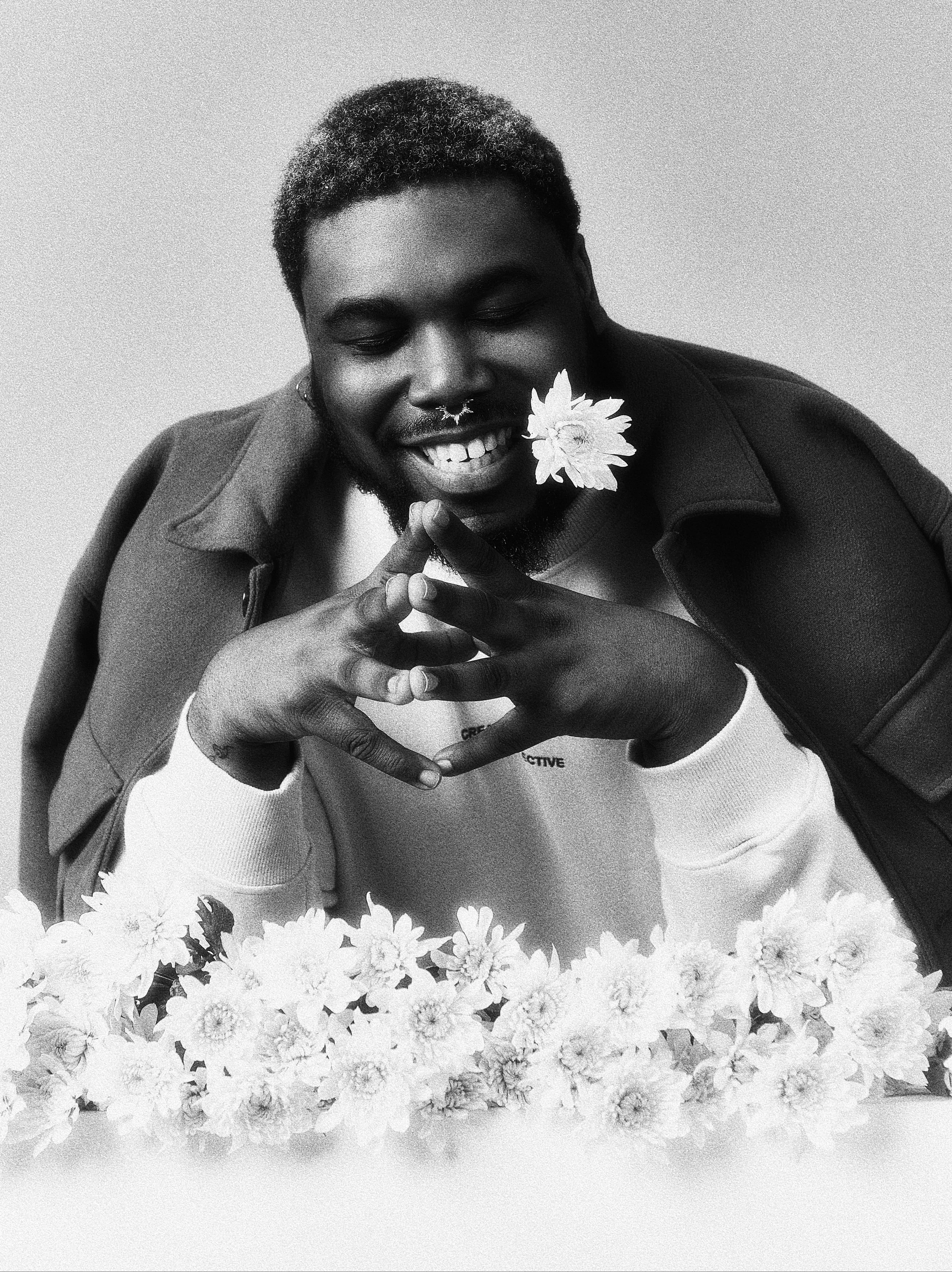Black and white photo of a smiling man with a flower in his mouth, hands clasped in front of him, surrounded by a border of flowers.