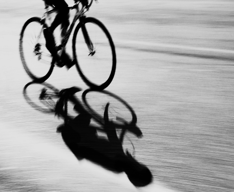A person riding a bicycle on wet pavement, with their reflection visible in the water.