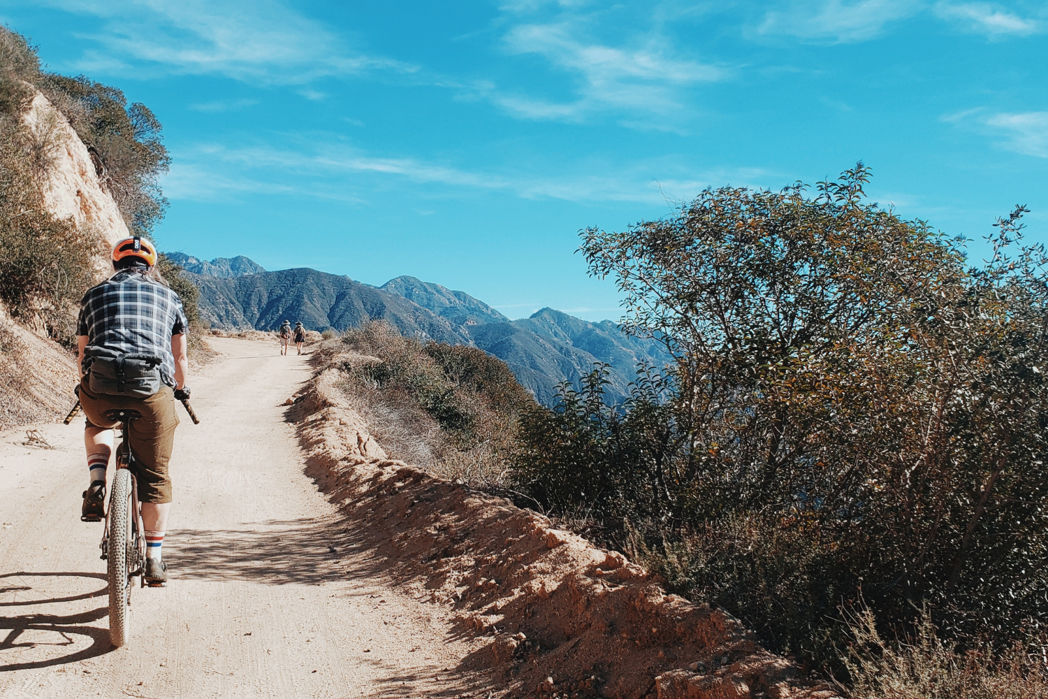 Person riding a mountain bike on a dirt trail with mountains and blue sky in the background.