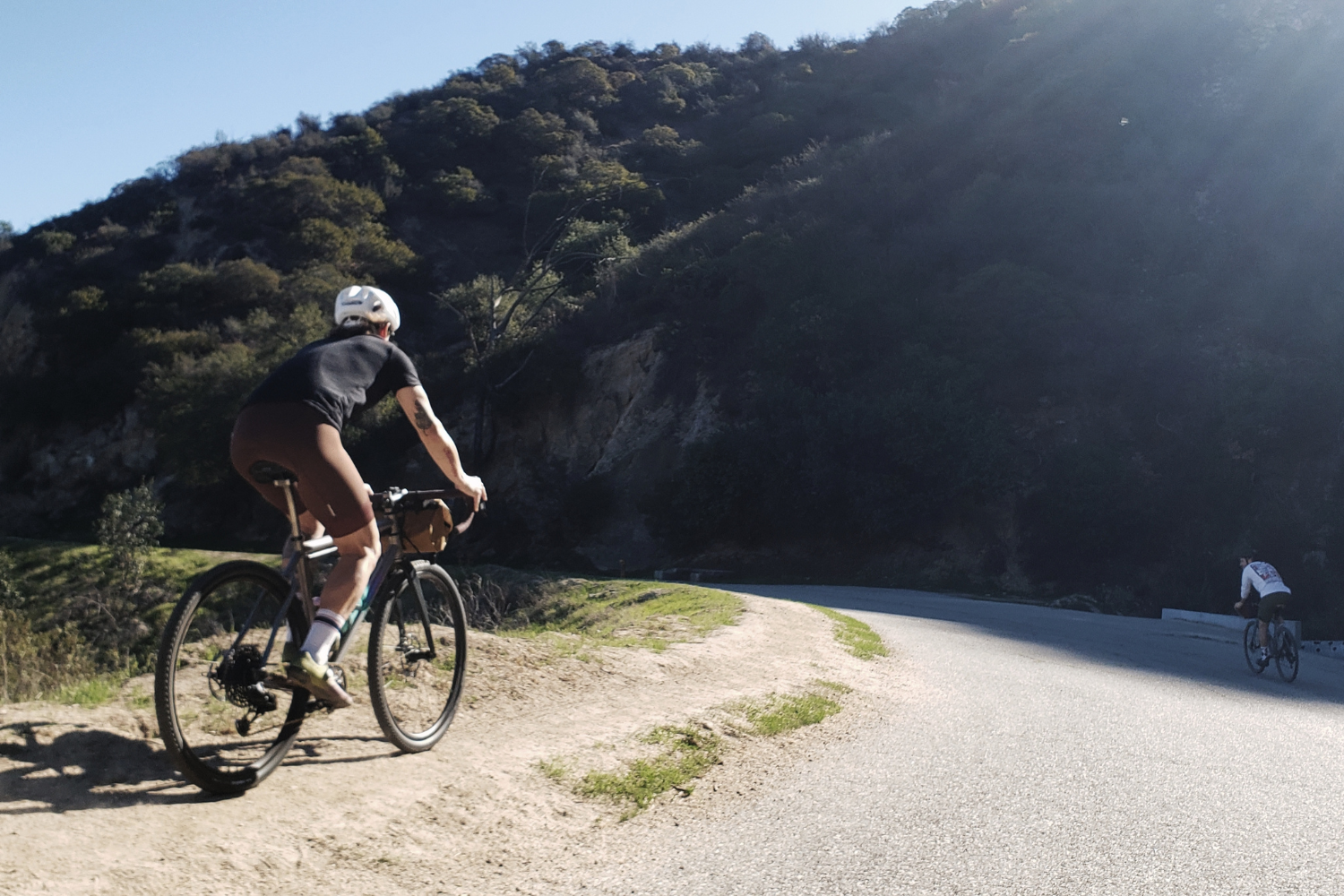 Two bikers riding on a winding mountain road with forested hills in the background on a sunny day.