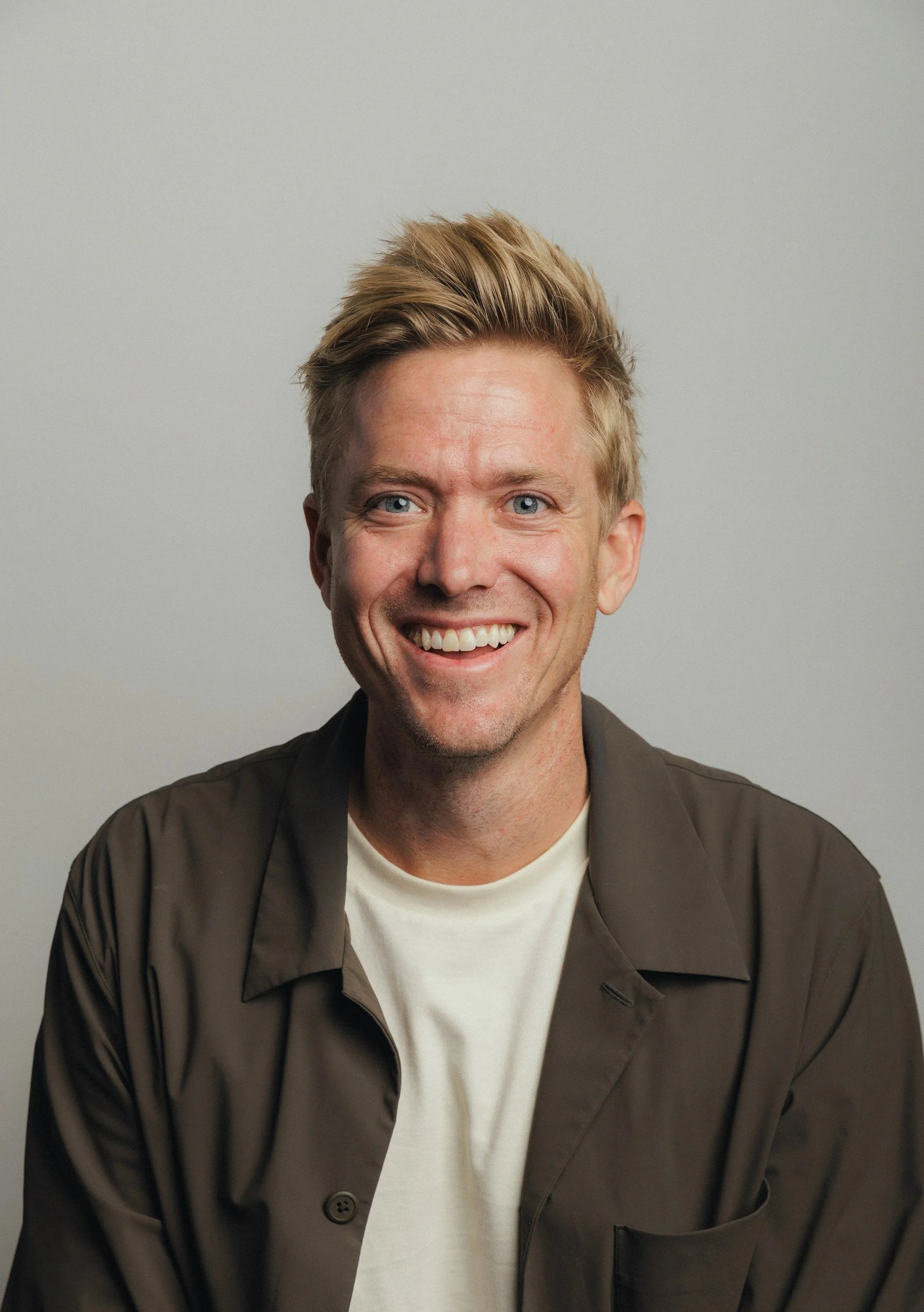 A young man with blonde hair and blue eyes smiling, wearing a white t-shirt and a dark brown jacket, against a plain light gray background.