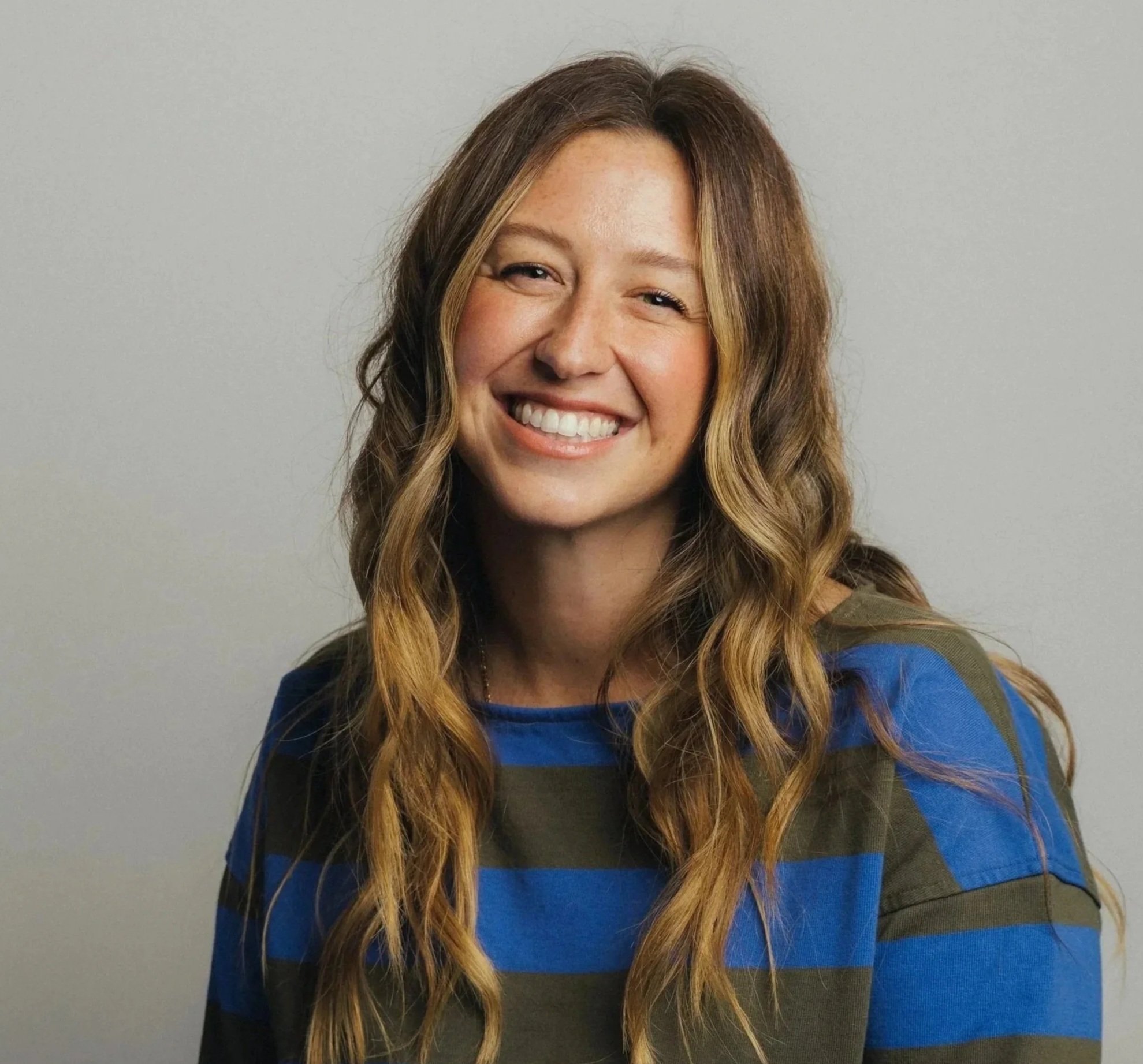 A smiling woman with long wavy hair and light skin, wearing a dark top with blue and gray striped sleeves, standing against a plain light gray background.