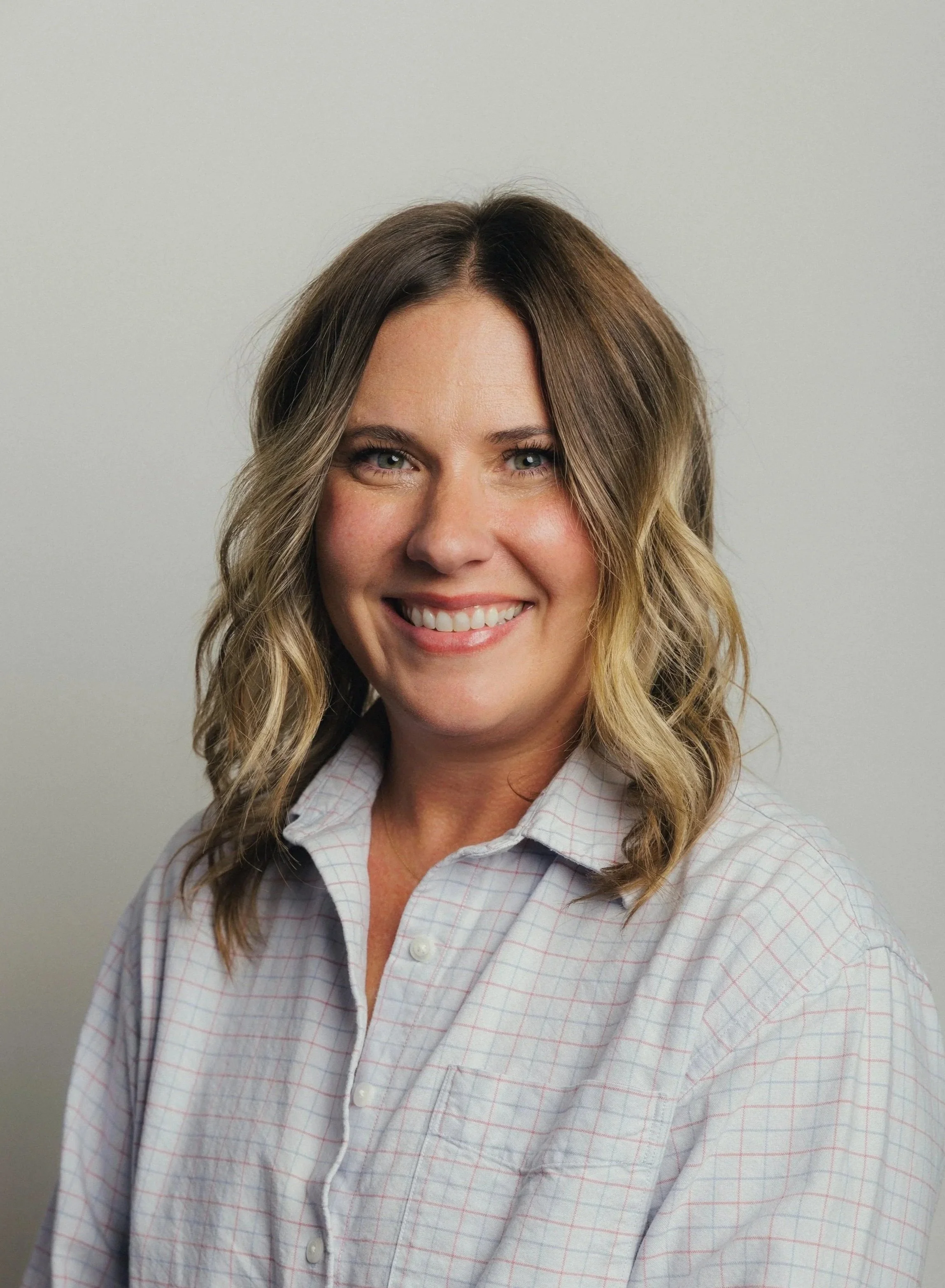 A woman with long, wavy, light brown hair with blonde highlights. She is smiling and wearing a light-colored, button-up shirt with a subtle grid pattern, standing against a plain, light-colored background.