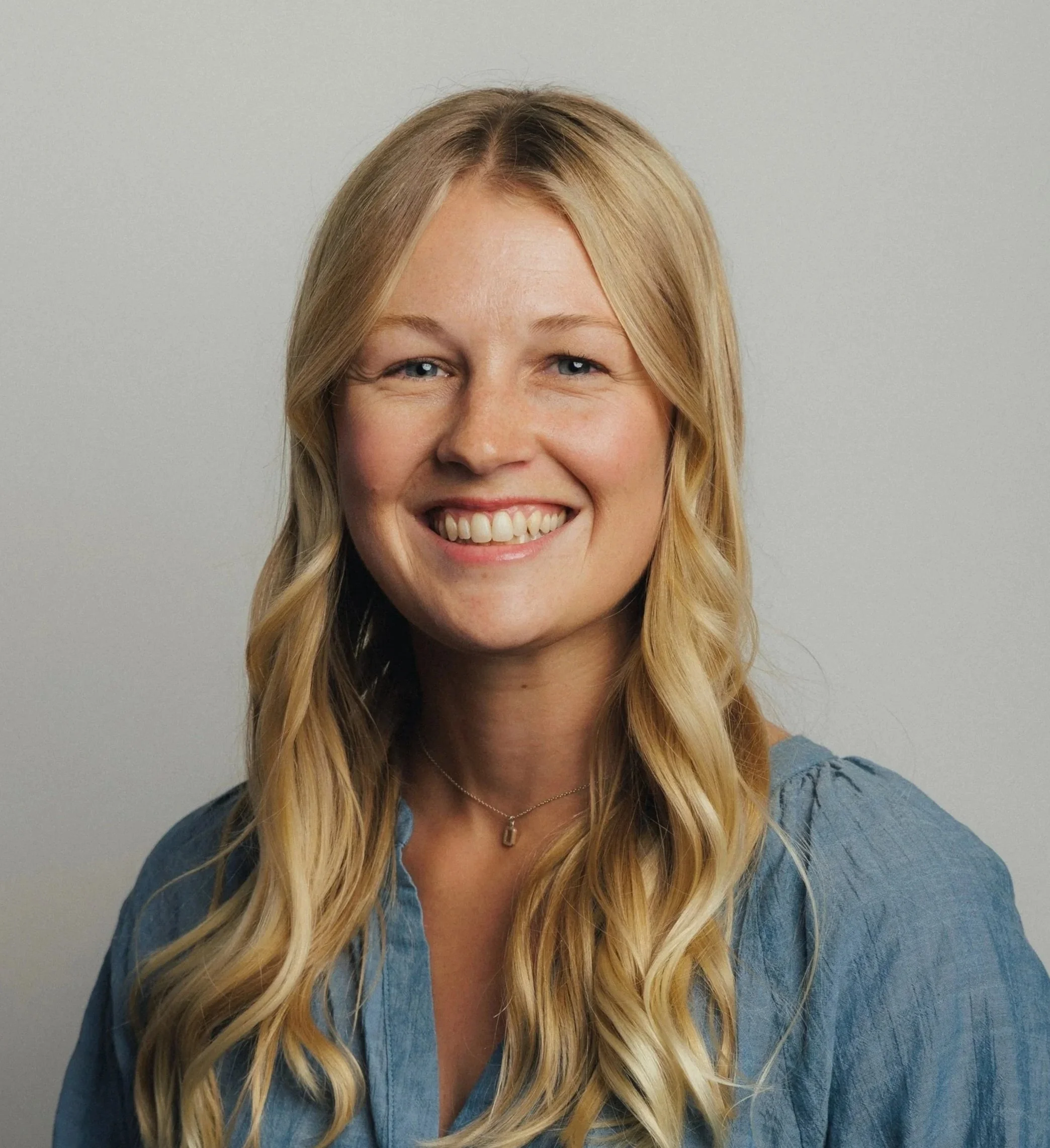 Headshot of a smiling woman with long wavy blond hair wearing a blue top and a silver necklace against a plain gray background.