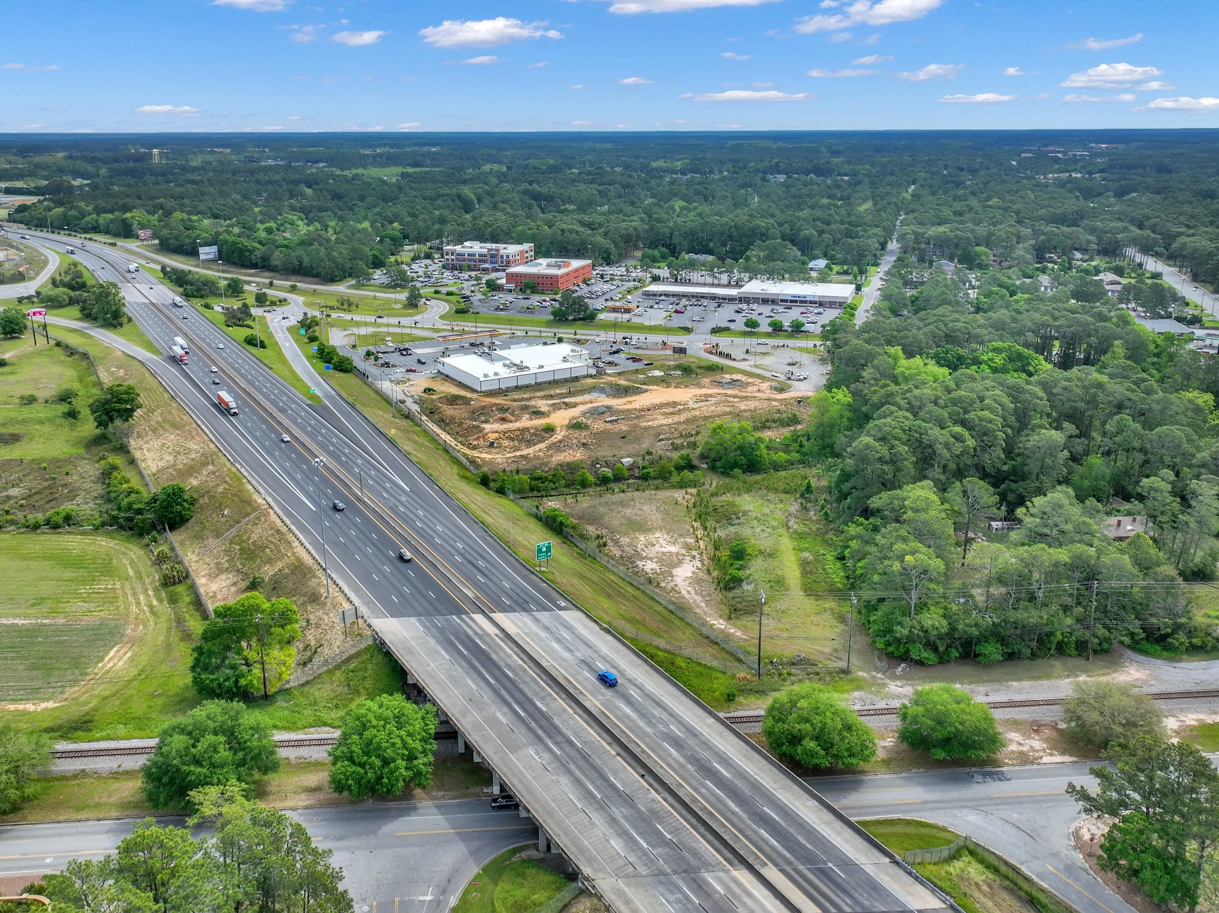 Aerial view of highways, commercial buildings, parking lots, and green trees under a partly cloudy sky.
