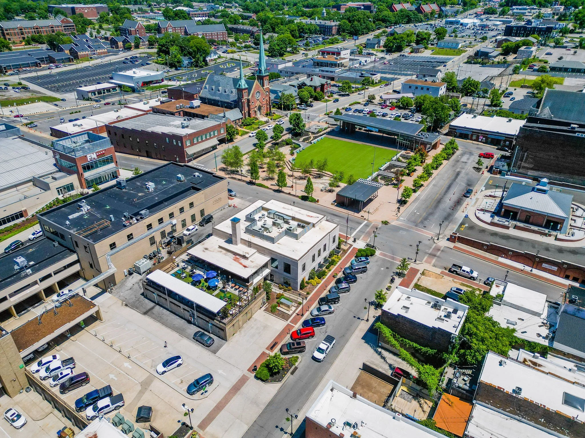 Aerial view of a cityscape showing a mix of buildings, parking lots, and green spaces, including a church with tall spires, a grassy park, and many parked cars.