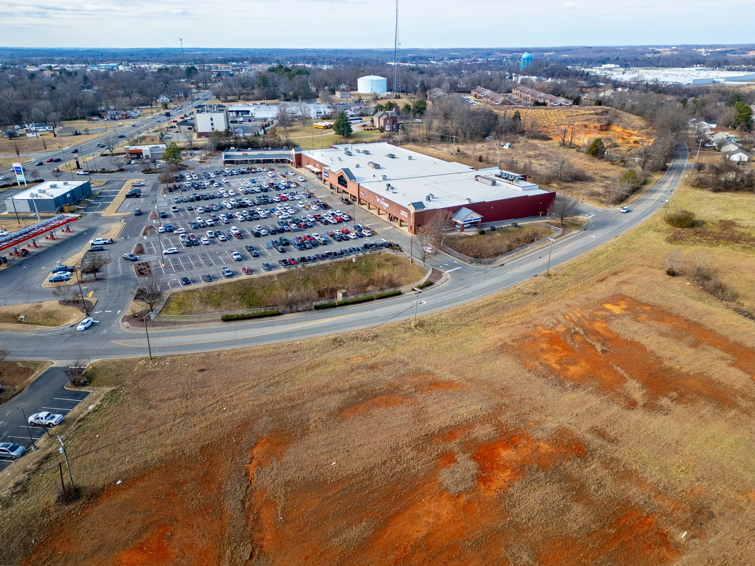Aerial view of a shopping center with a large parking lot surrounded by roads, with empty and occupied parking spaces. The shopping center has a red and white facade and a nearby gas station. There are open land and some trees around, with dirt patch
