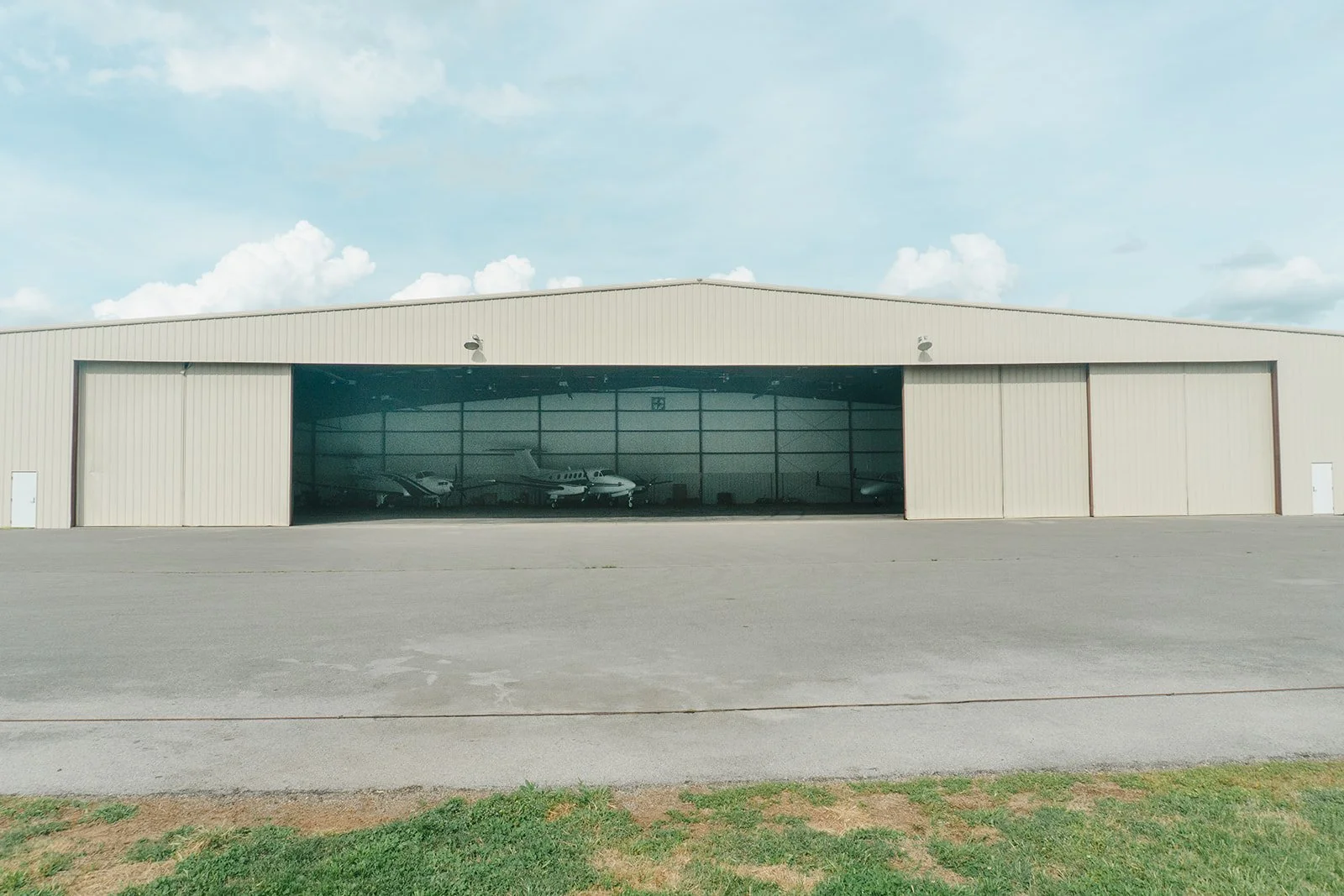 Large airplane hangar with a partially open door showing several small aircraft inside, set on a concrete apron with grass in the foreground under a partly cloudy sky.