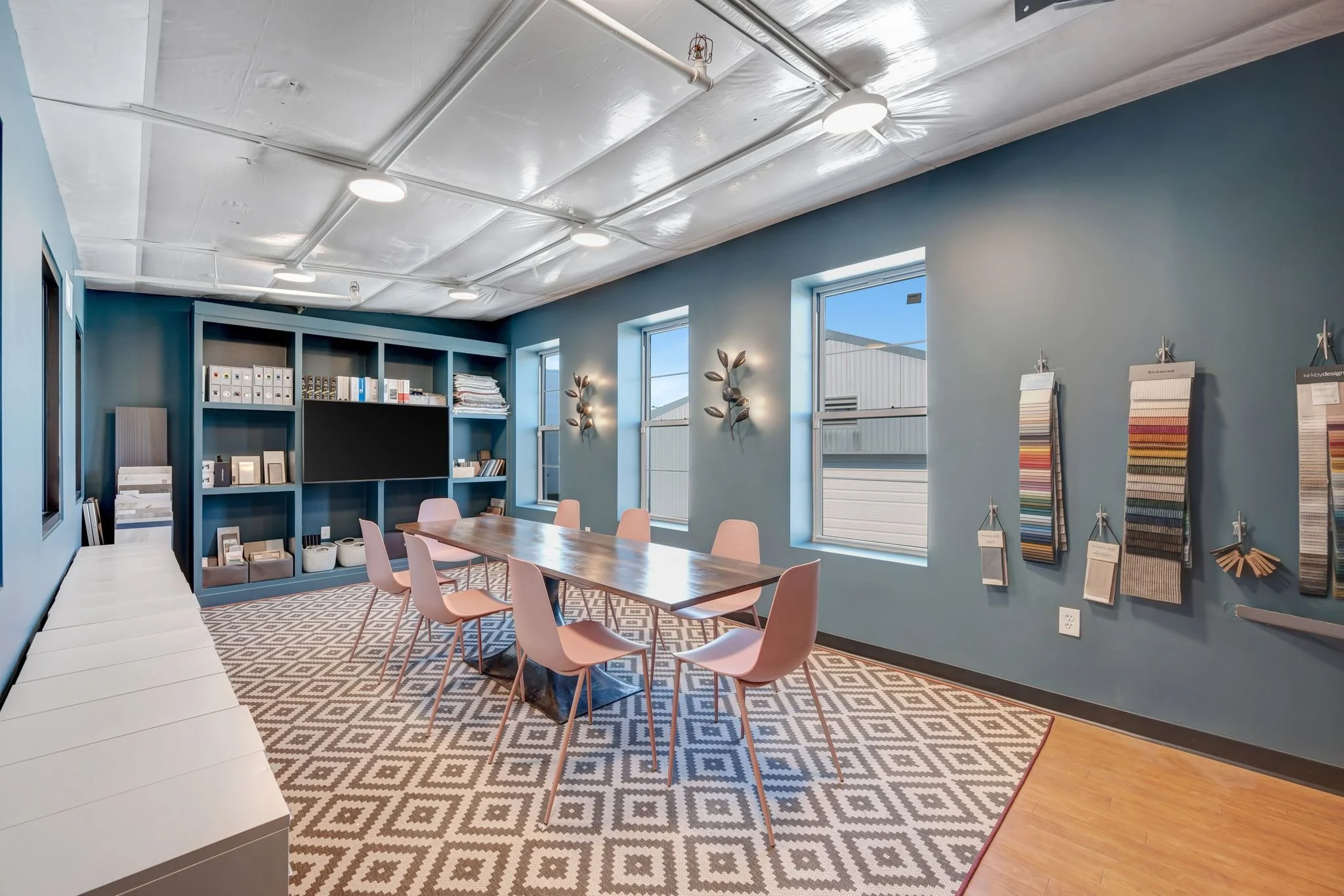 A modern meeting room with a long wooden table, pink chairs, a patterned rug, and a blue wall with shelves, windows, and decorative wall art.
