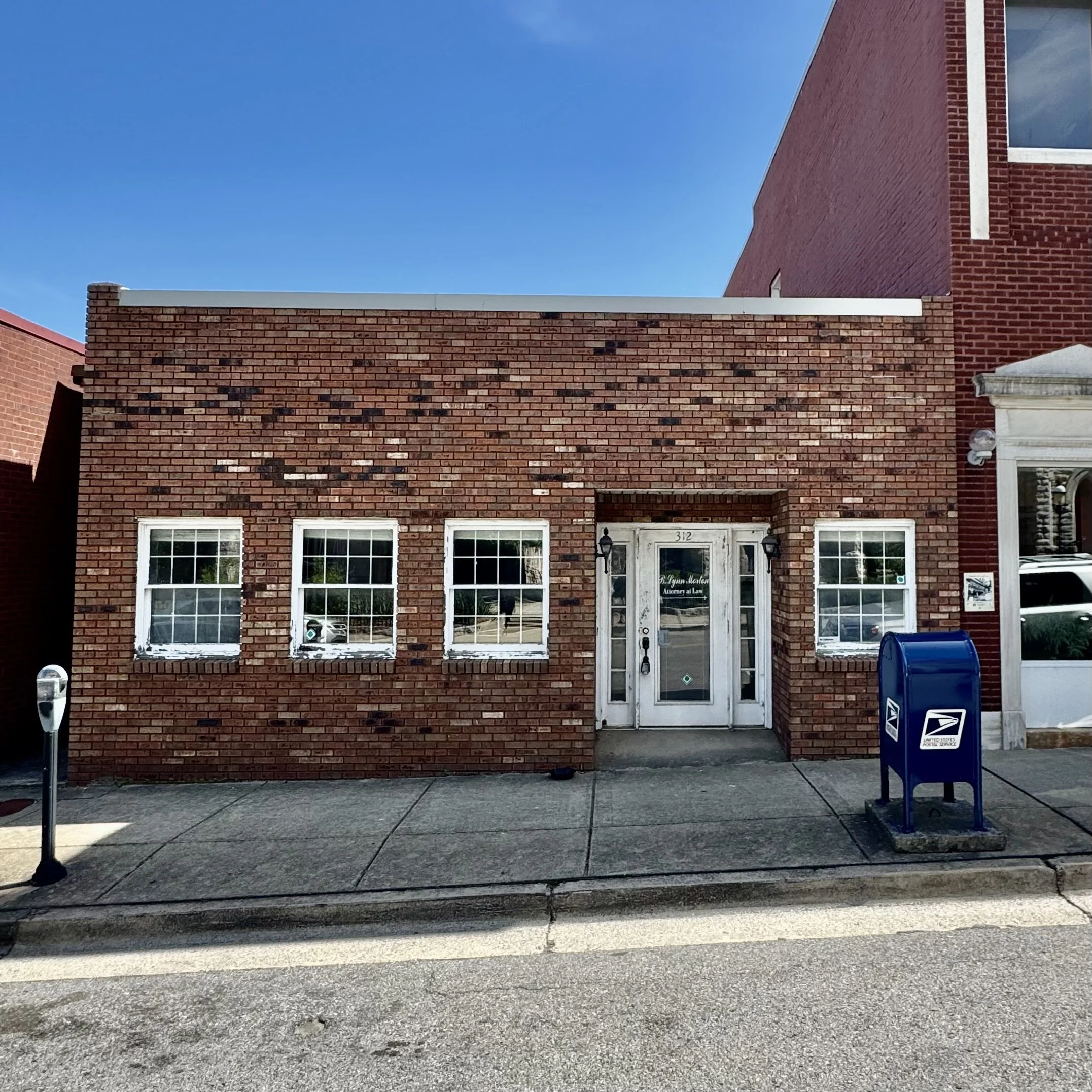 A small brick building with three windows and a glass door, located on a sidewalk with a blue USPS mailbox in front.